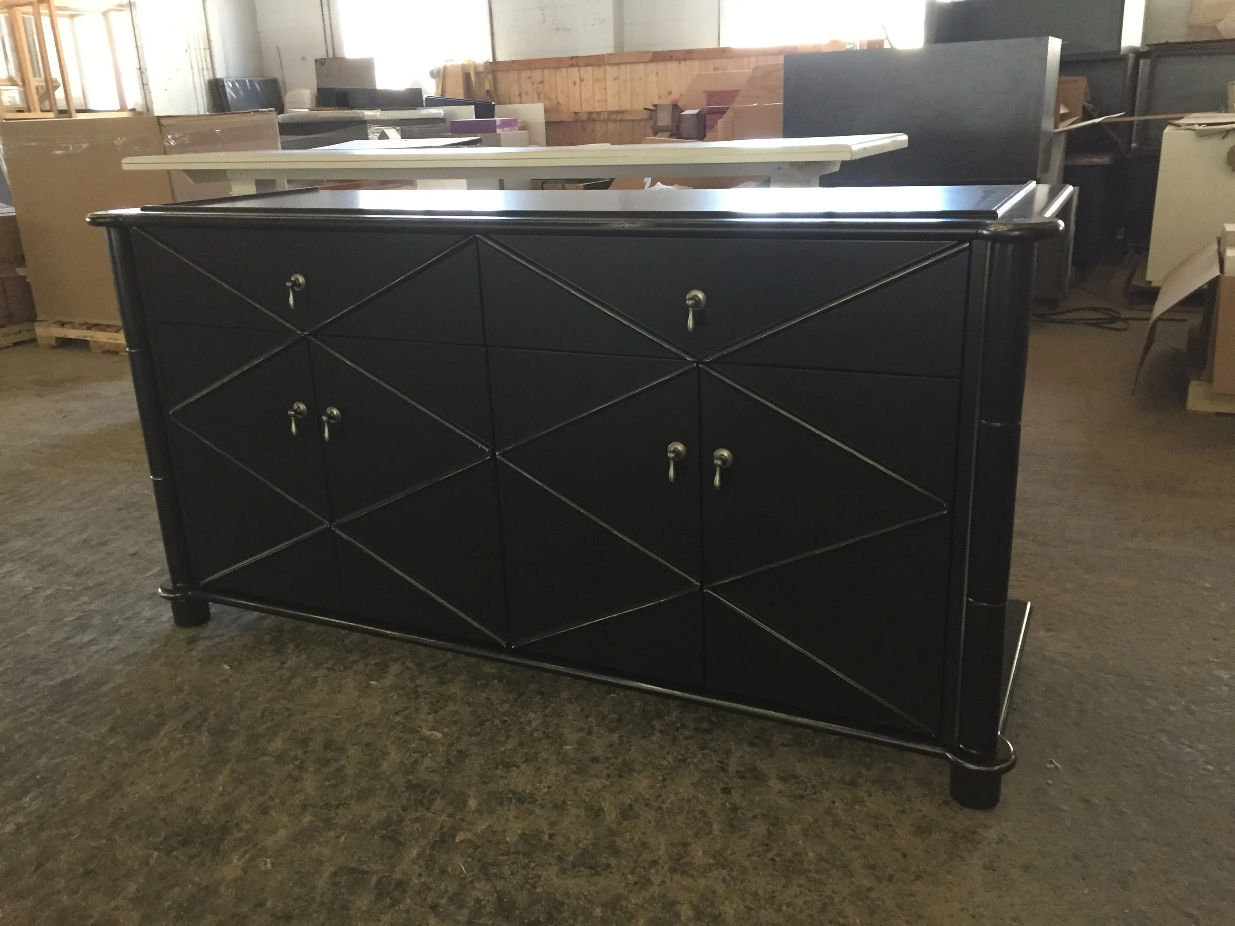 A black dresser with silver knobs, standing on a concrete floor in a warehouse or storage space with various furniture pieces and boxes in the background.