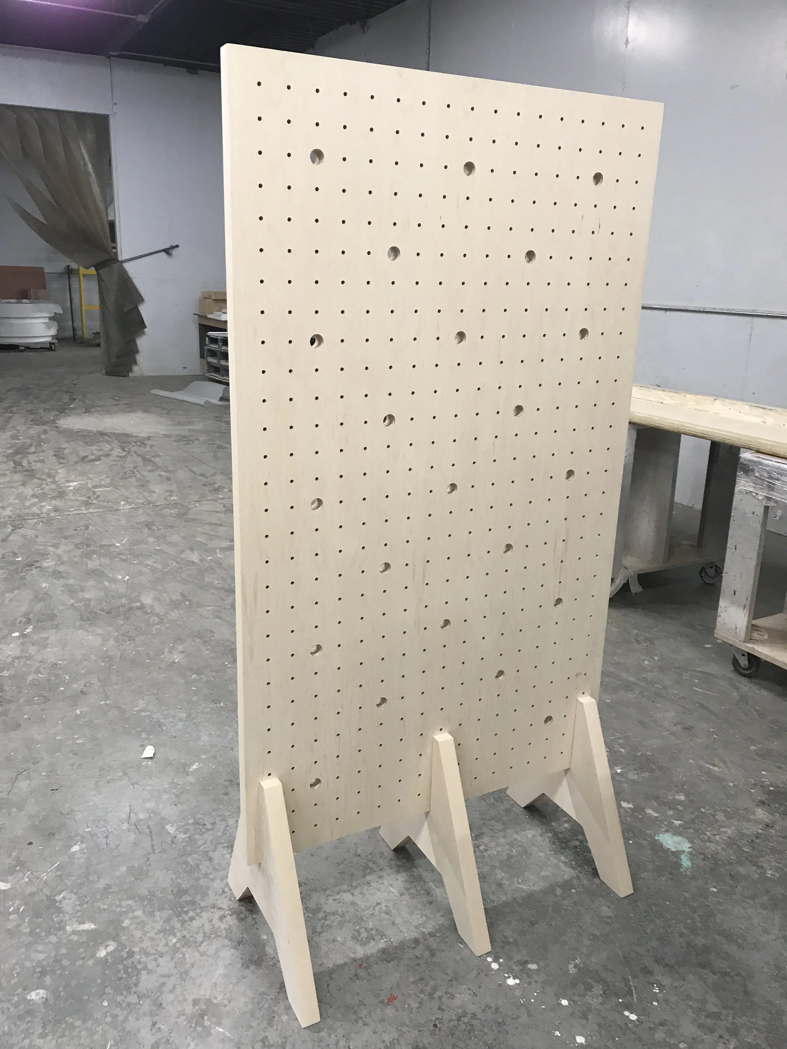 A wooden display board standing in a workshop with a pegboard design, four holes visible, and a beige color, supported by wooden legs.