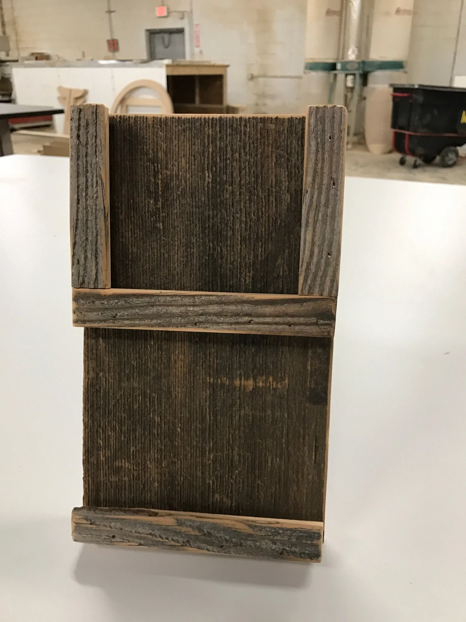 Close-up of a small wooden box with weathered wood grain, placed on a white surface in a workshop or construction area with tools and equipment in the background.