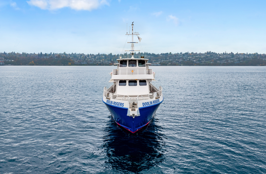 A boat named Doolin-Rogers is on the water, facing directly towards the camera, with land and trees visible in the background under a blue sky.