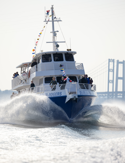 A boat moving swiftly on the water with a bridge in the background, wearing nautical decorations and carrying several people.