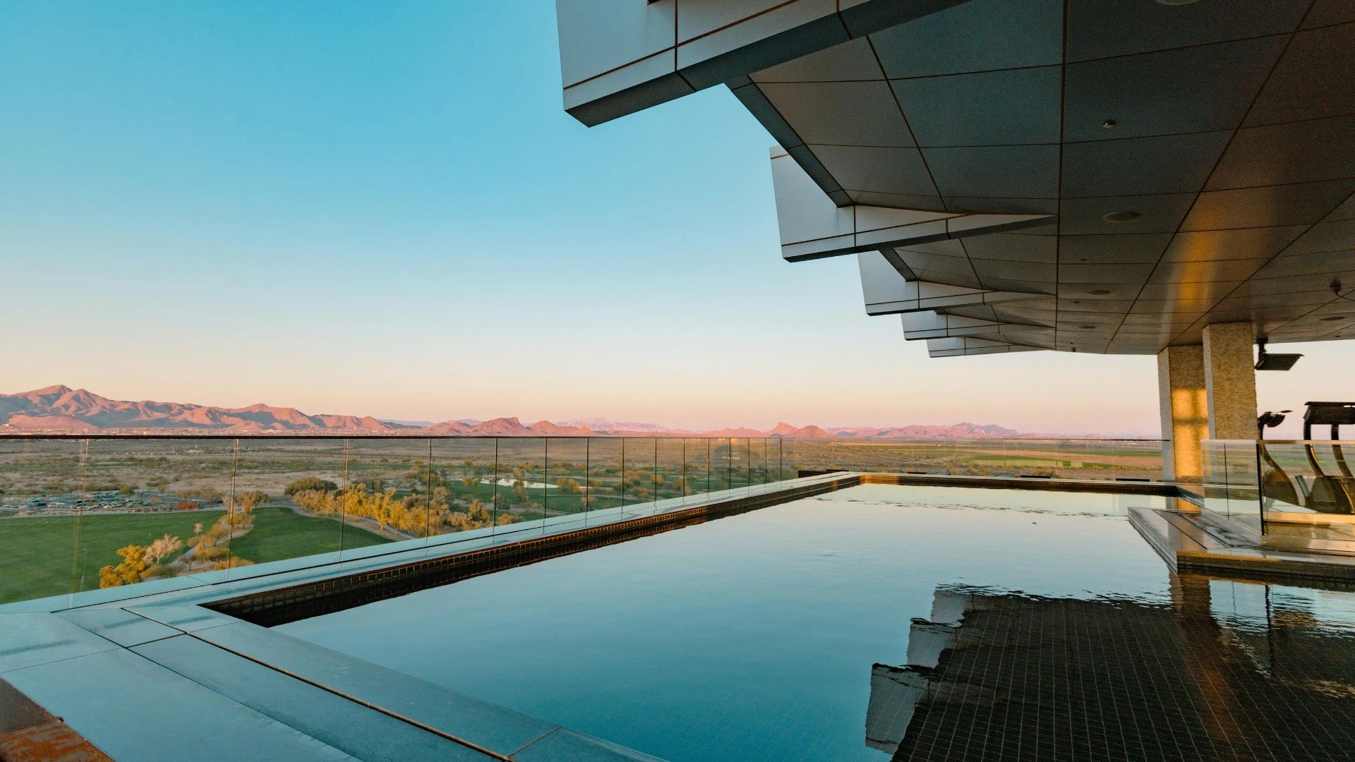 View of a modern building with an infinity pool on a balcony, overlooking a landscape with mountains, fields, and a clear sky at sunset.