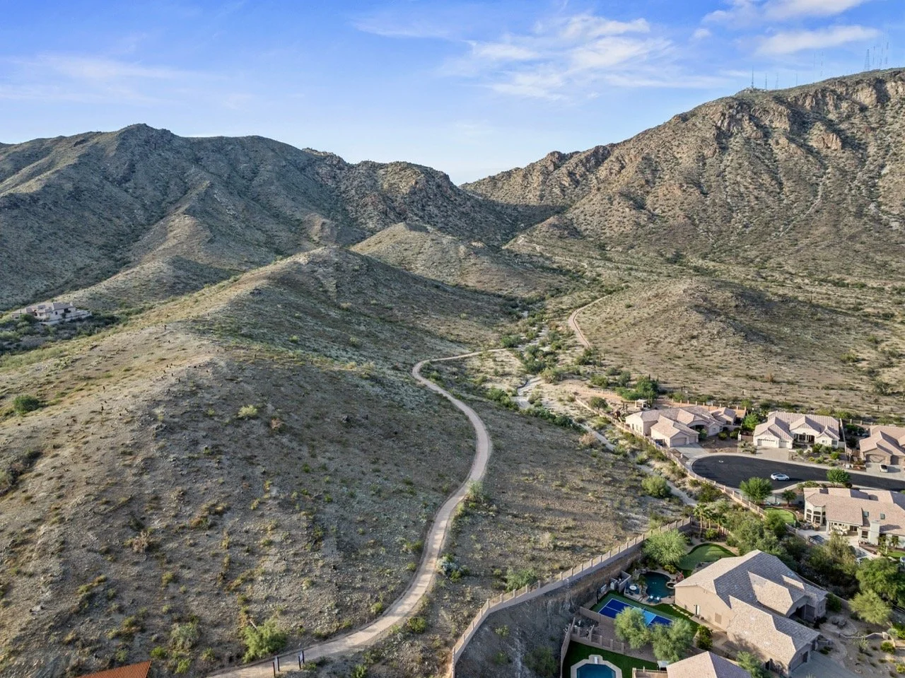 A winding trail runs through a dry, hilly desert landscape with sparse vegetation and residential homes in the foreground. Mountains are visible in the background under a partly cloudy sky.