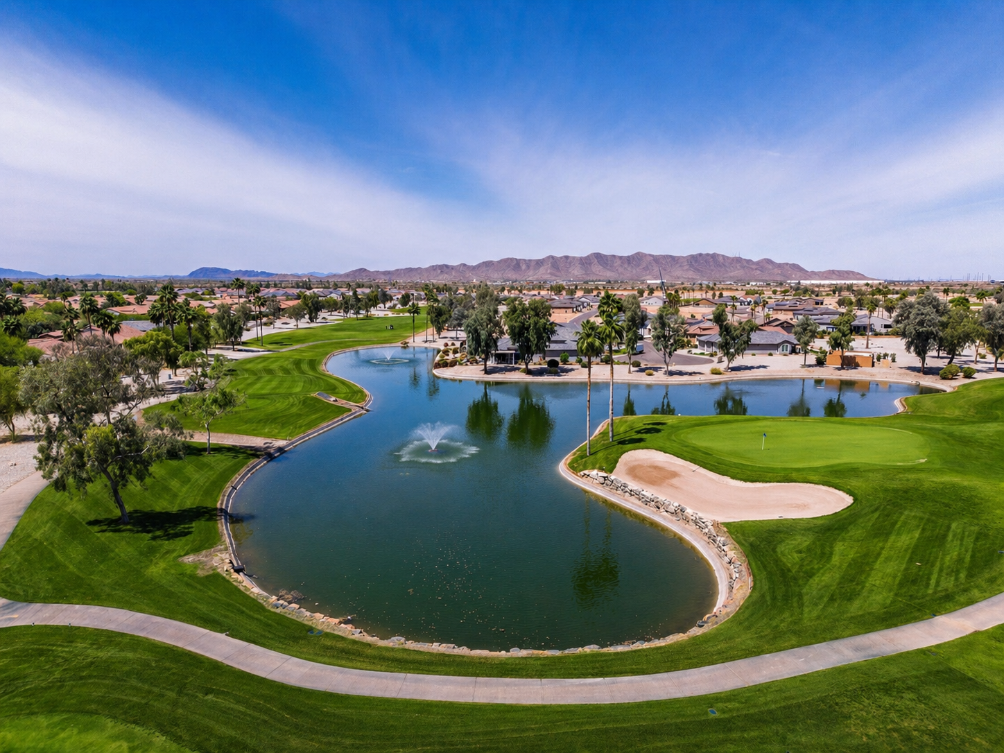 Aerial view of a golf course with a pond, fountains, green grass, trees, and adjacent houses with a mountain range in the background.