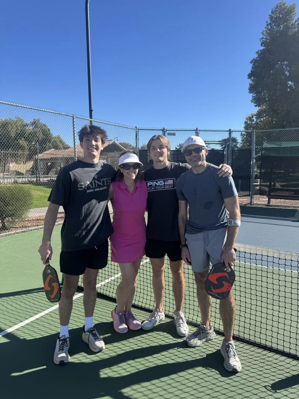 Four people standing together on a tennis court, holding pickleball paddles, posing for a photo on a sunny day.