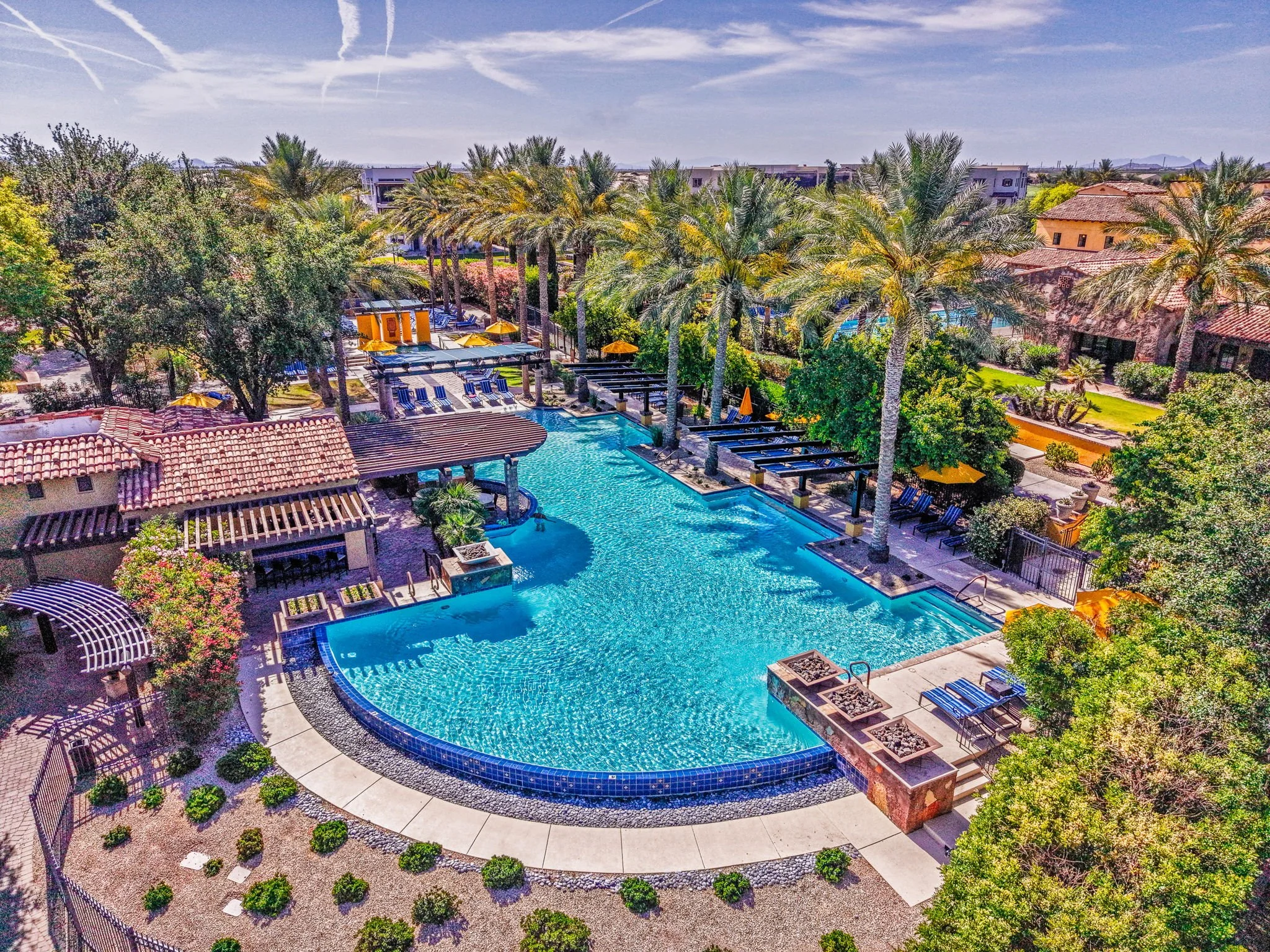 An aerial view of a luxurious swimming pool area at a resort surrounded by palm trees, lounge chairs, and cabins, with a clear blue sky overhead.