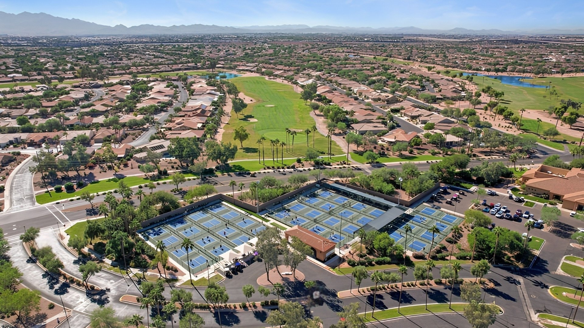 Aerial view of a suburban area featuring houses, a park with green lawns, trees, a pond, and a tennis court complex with solar panels, with mountains in the background.