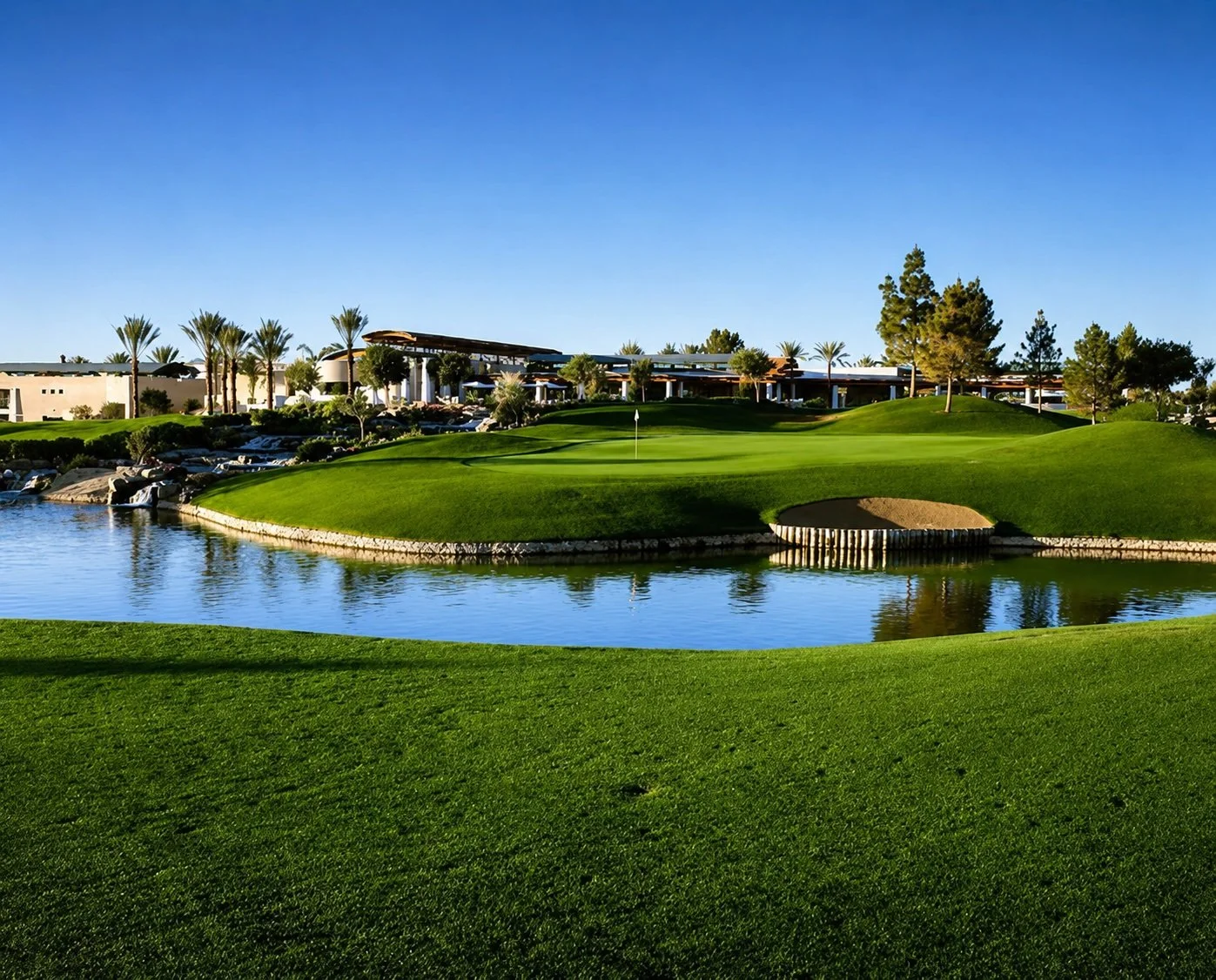 A golf course with a green surrounded by water, sand bunkers, and lush grass, with a clubhouse and palm trees in the background under a clear blue sky.