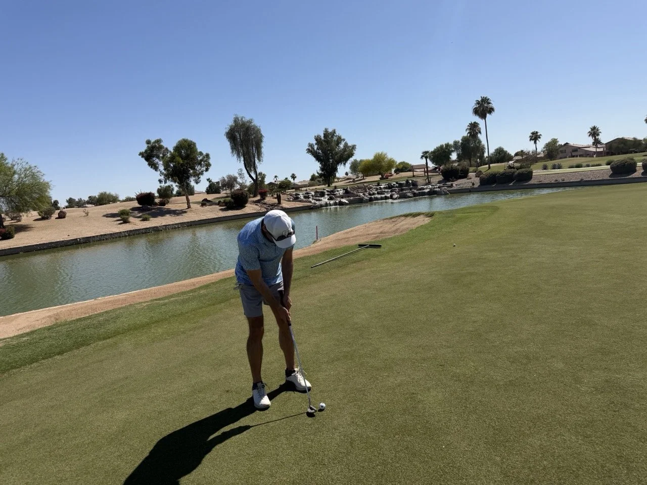 Man preparing to hit a golf ball on a putting green near a water hazard at a golf course, with desert landscape and palm trees in the background on a sunny day.