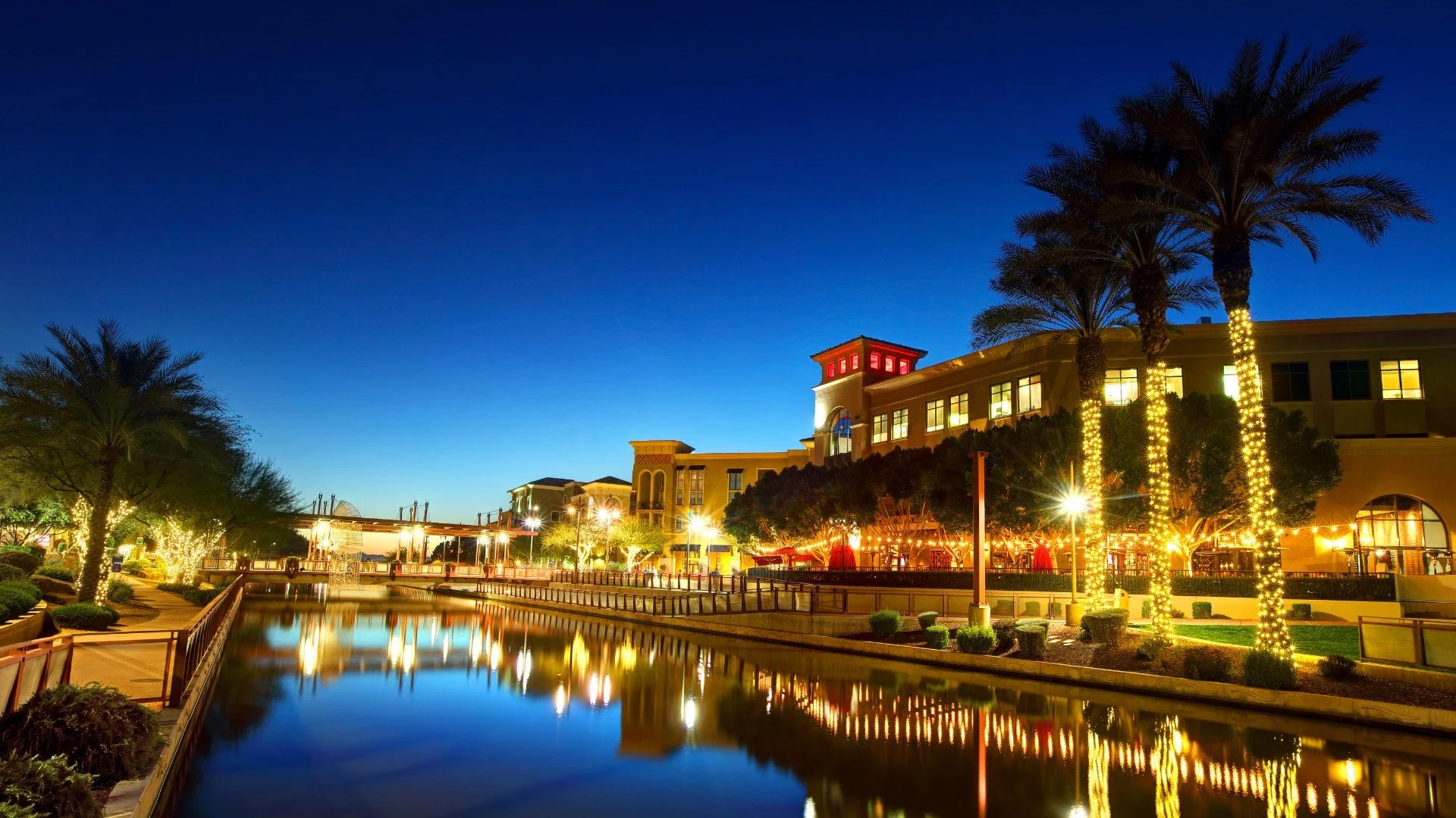 A cityscape at dusk with a building illuminated by warm lights, palm trees wrapped in string lights, a waterway reflecting the lights, and a clear sky transitioning from deep blue to lighter shades near the horizon.