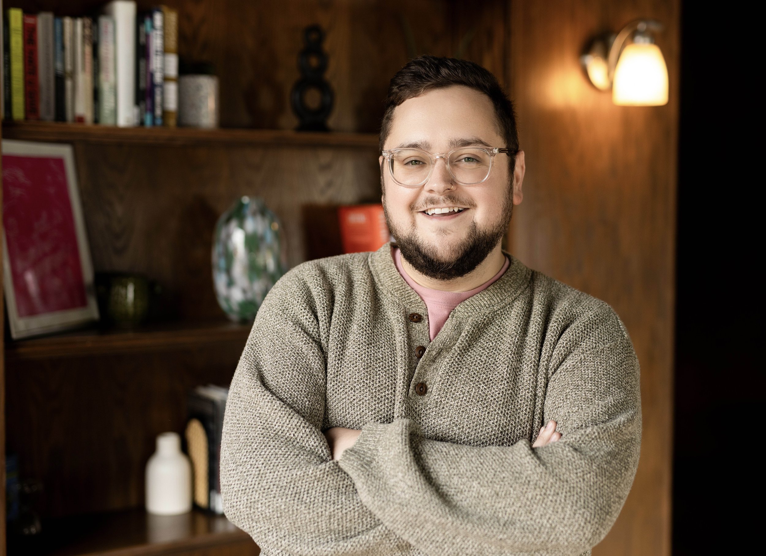 Smiling therapist standing with arms crossed in a warm, cozy office. He is wearing glasses and a textured neutral sweater, with bookshelves, soft lighting, and wood-paneled walls in the background.