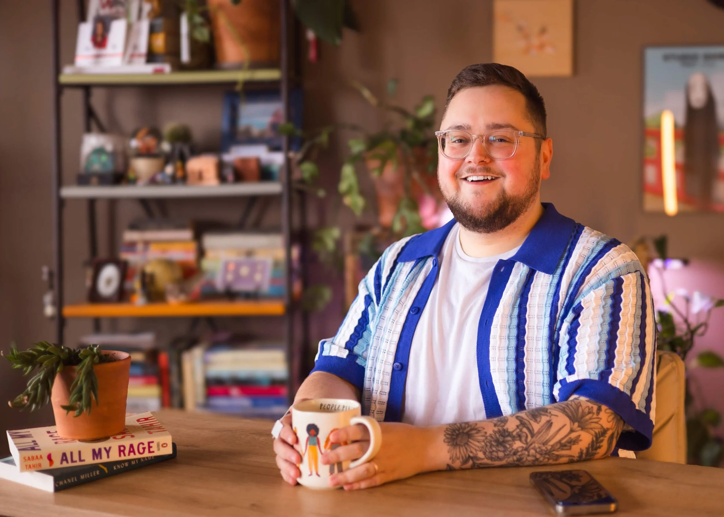 Kody Garis smiling while seated at a wooden desk in a warm, cozy office. He is wearing glasses, a white shirt under a blue-and-white textured button-up shirt, with floral tattoos visible on his left forearm. He is holding a mug with books in the back