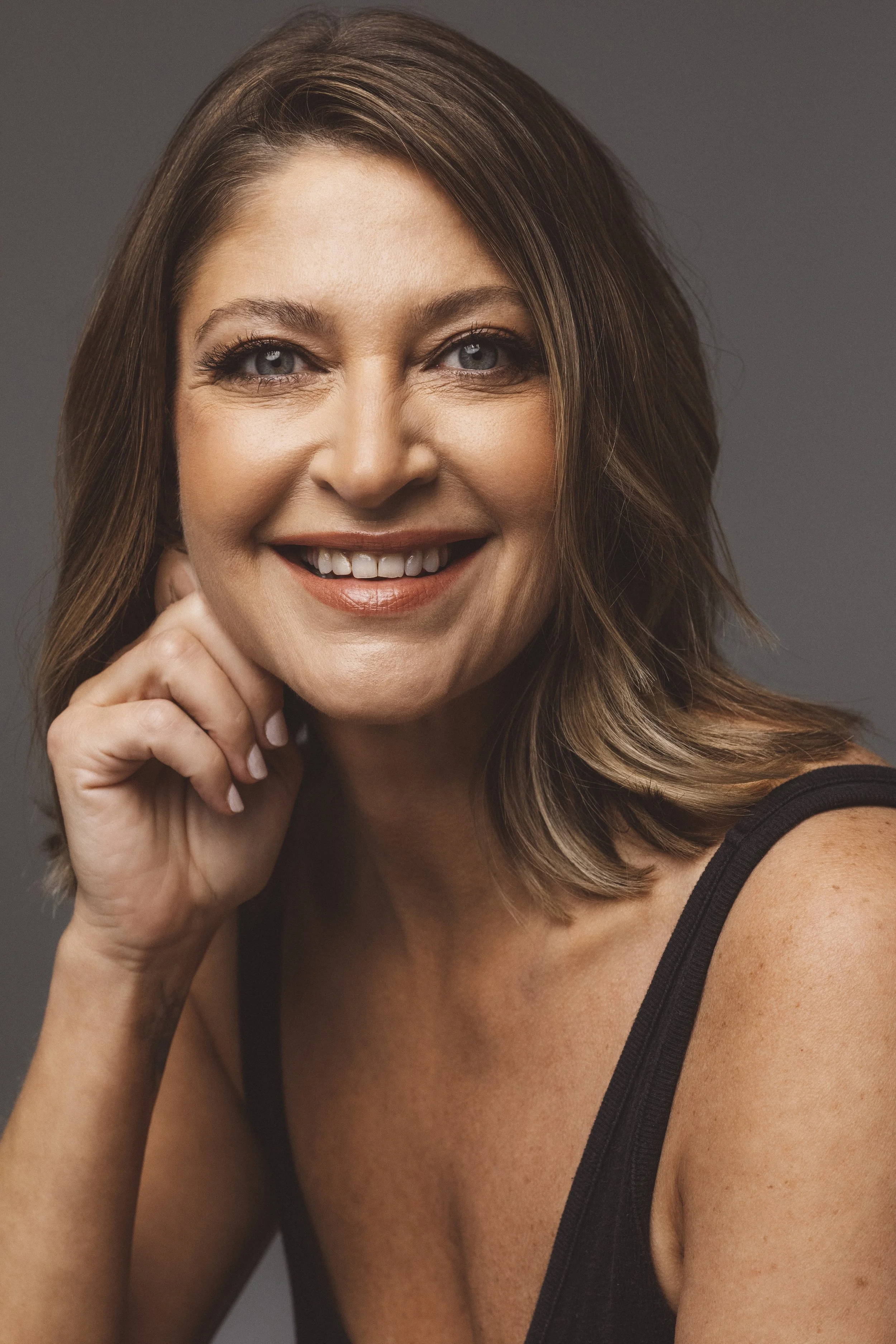 Close-up portrait of a smiling woman with blue eyes and shoulder-length brown hair, wearing a black tank top, against a gray background.