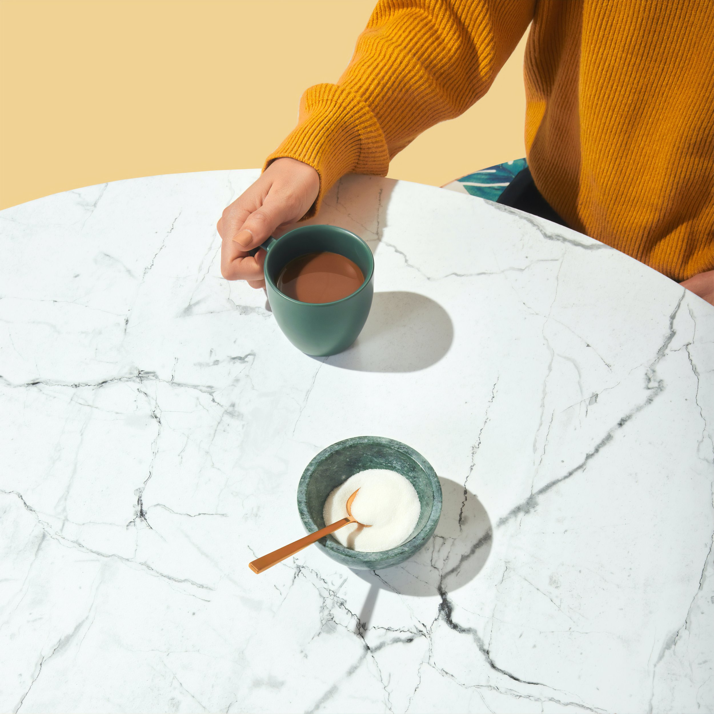 A hand holding a green mug filled with hot chocolate on a white marble table with black veining. The background is a solid beige color, and a spoon is in the mug.