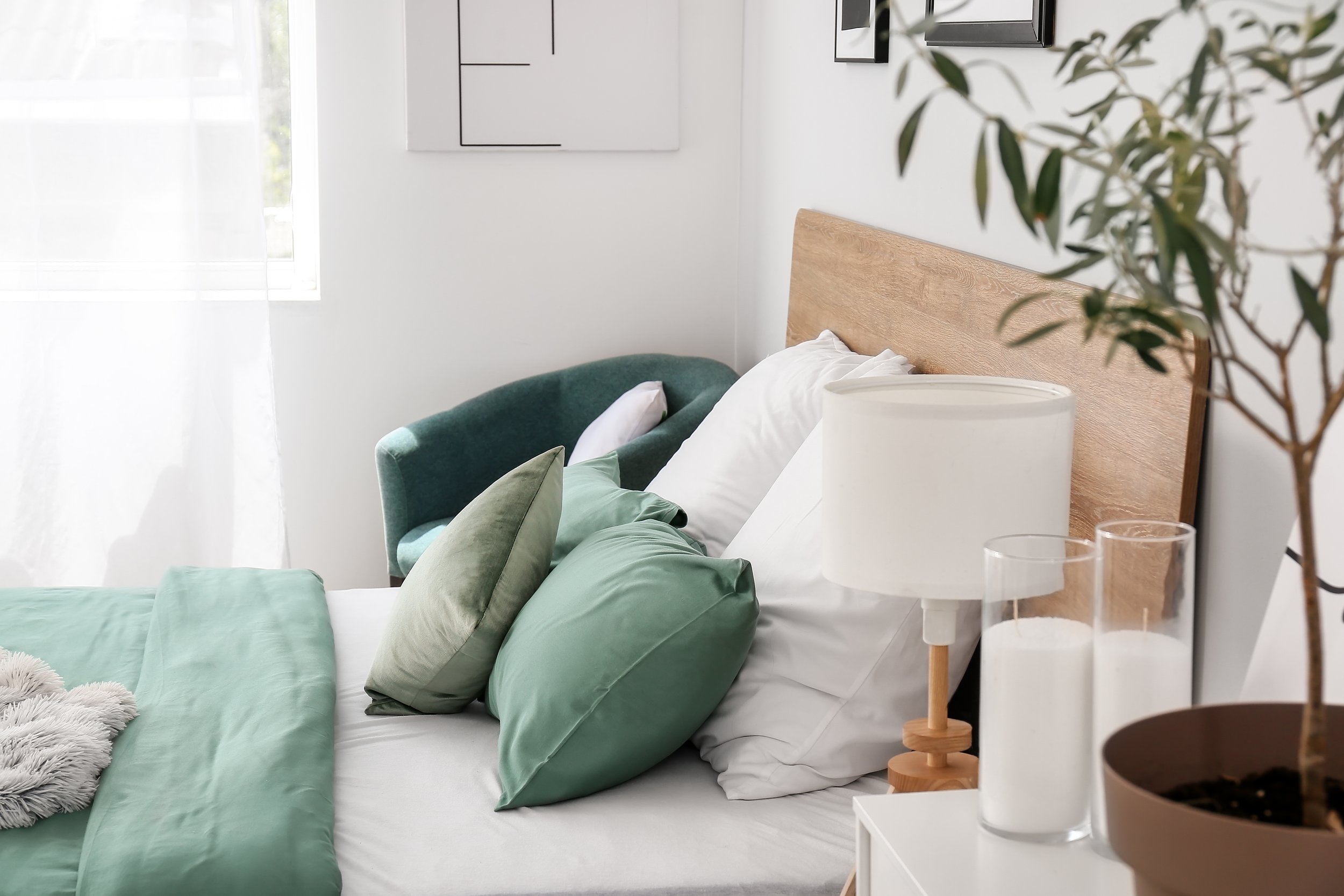 A cozy bedroom with a large bed featuring green and white pillows, a wooden headboard, a white nightstand with a lamp, and a window with sheer curtains letting in natural light. There are framed pictures on the wall and a leafy plant to the right.
