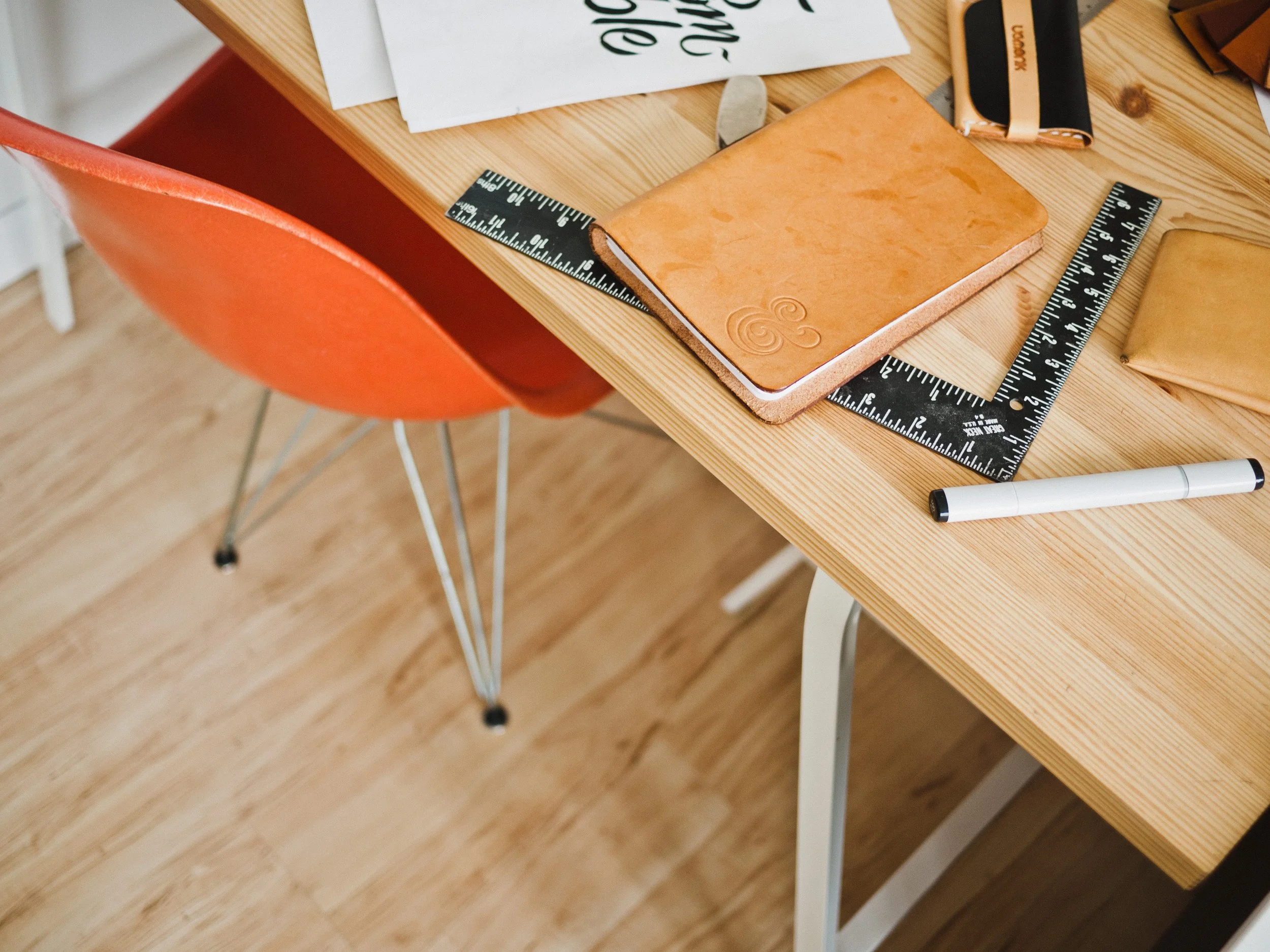 A wooden desk with a notebook, black and orange item, ruler, a white marker, and some papers, next to an orange chair with thin metal legs.