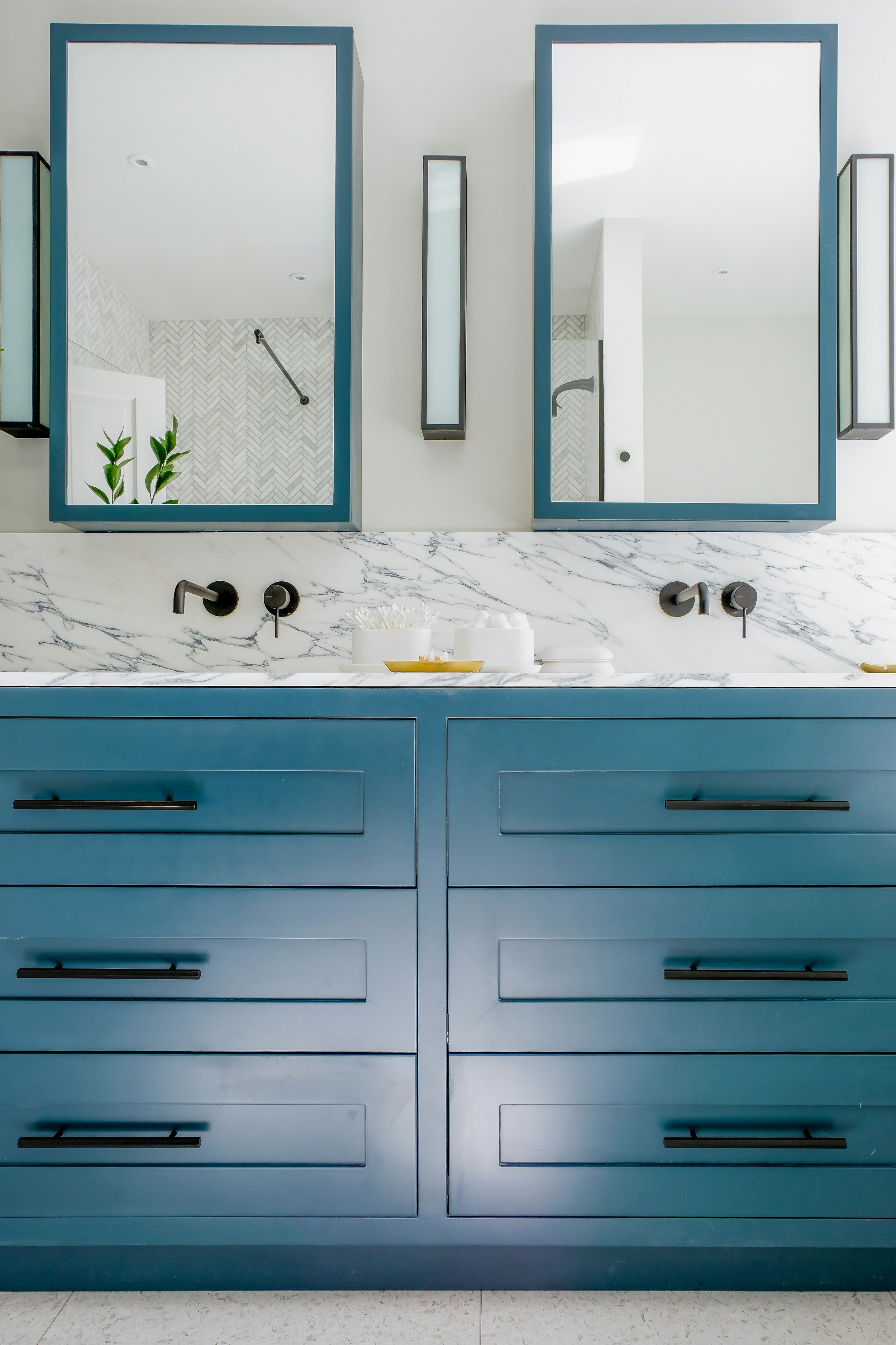 Modern bathroom vanity with blue cabinets, marble countertop, and large mirror with black frames, reflecting a shower area with a herringbone tile wall and plants.
