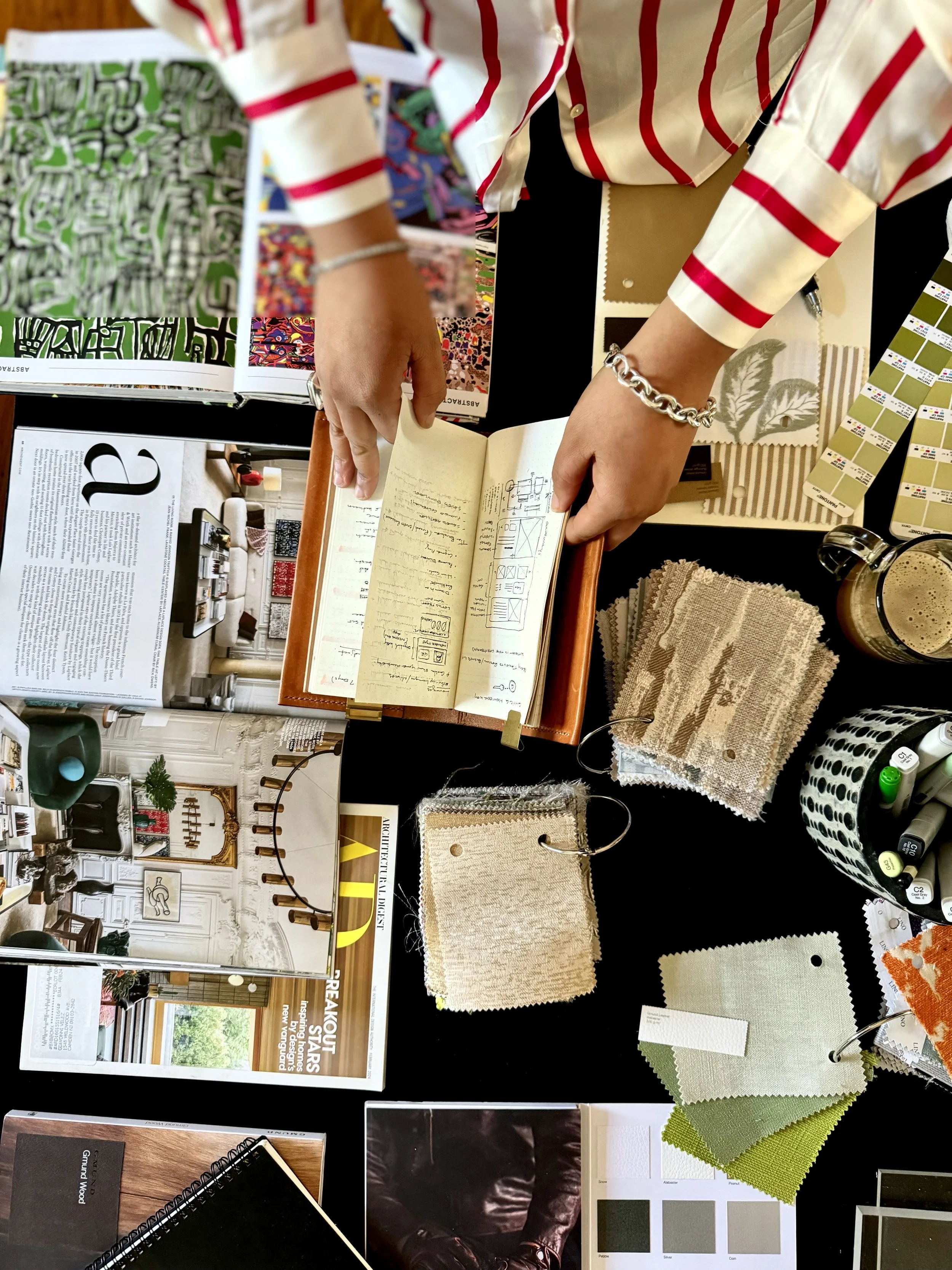 A person wearing a white shirt with red stripes is standing at a table with various fabric samples, color swatches, and a notebook. The table has magazine pages, paint chips, and fabric swatches in different textures and patterns.