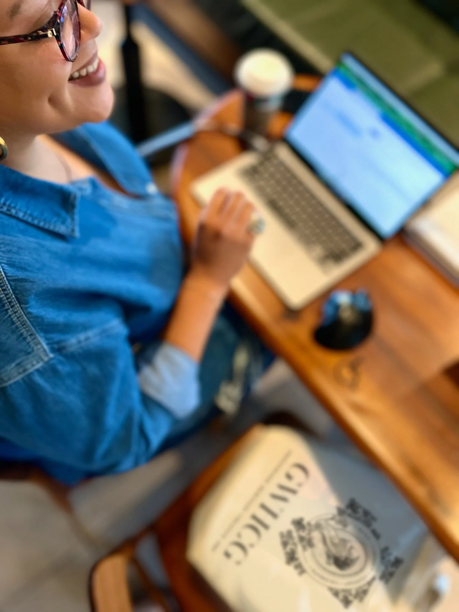 A woman wearing glasses sitting at a wooden table with a laptop and a drink, smiling while looking at her laptop screen.