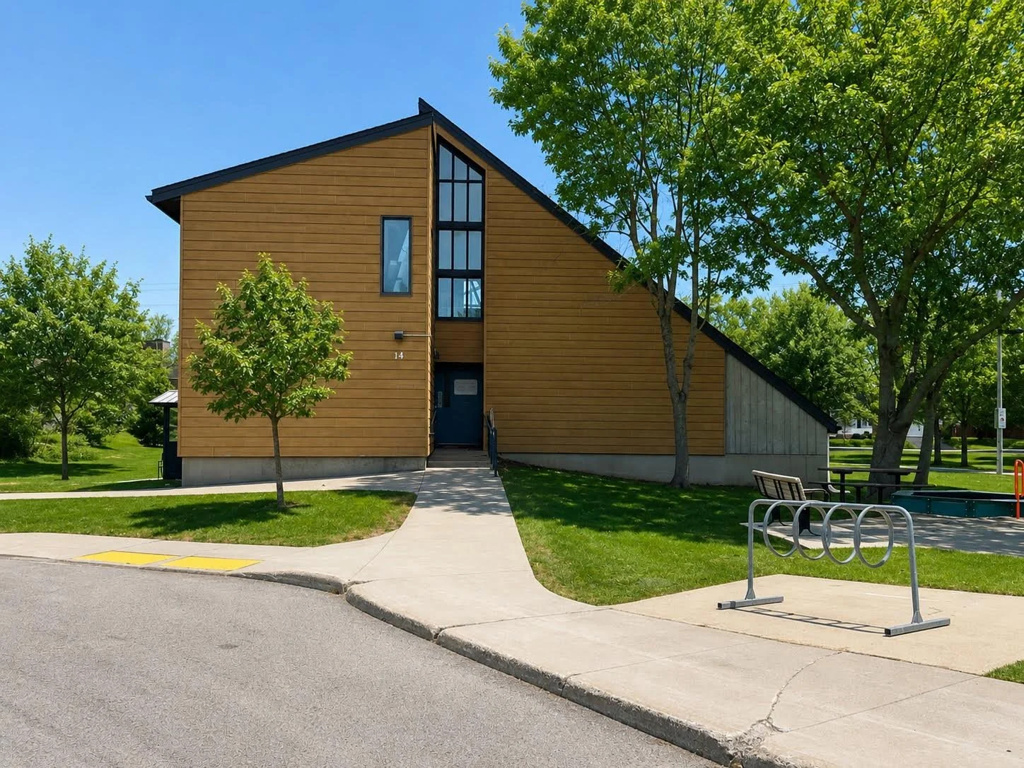 Modern wooden building with stairs at the entrance, surrounded by green trees and grass, with a bike rack in the foreground.