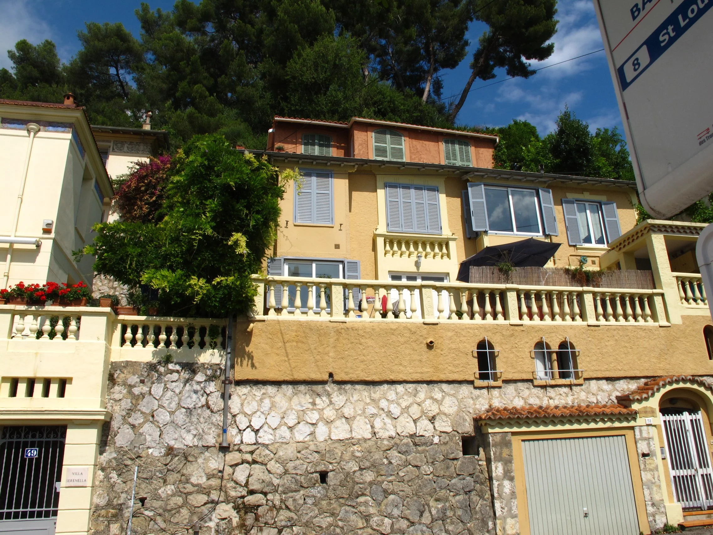 Multi-story houses with pastel-colored facades and shutters, situated on a hillside with lush green trees in the background, under a blue sky with some clouds.