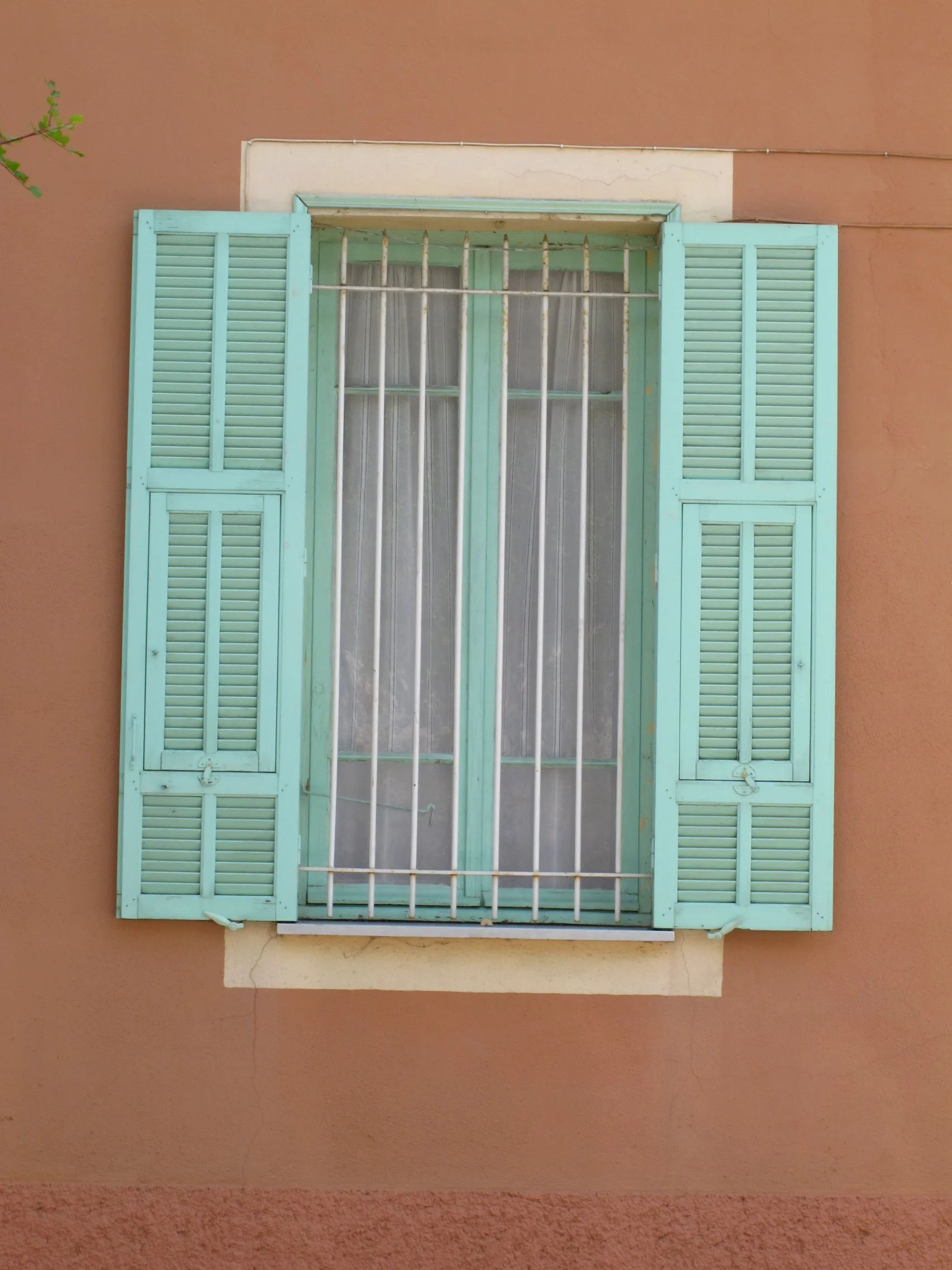 A window with closed light blue shutters and iron security bars, set into a beige wall.