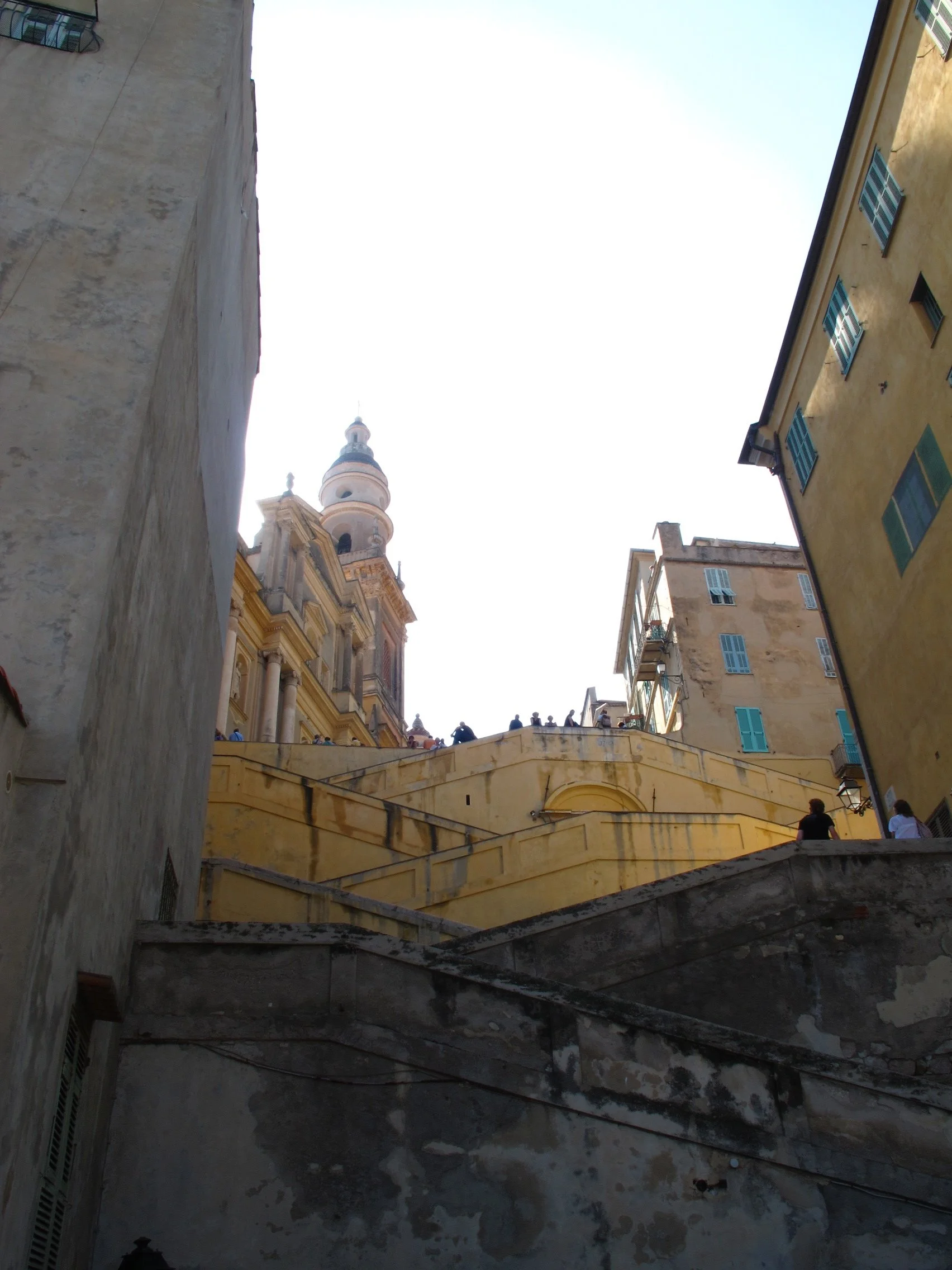 A narrow alleyway between old buildings with stairs leading up to a terrace where people are gathered, with a church tower visible in the background, in a European city.