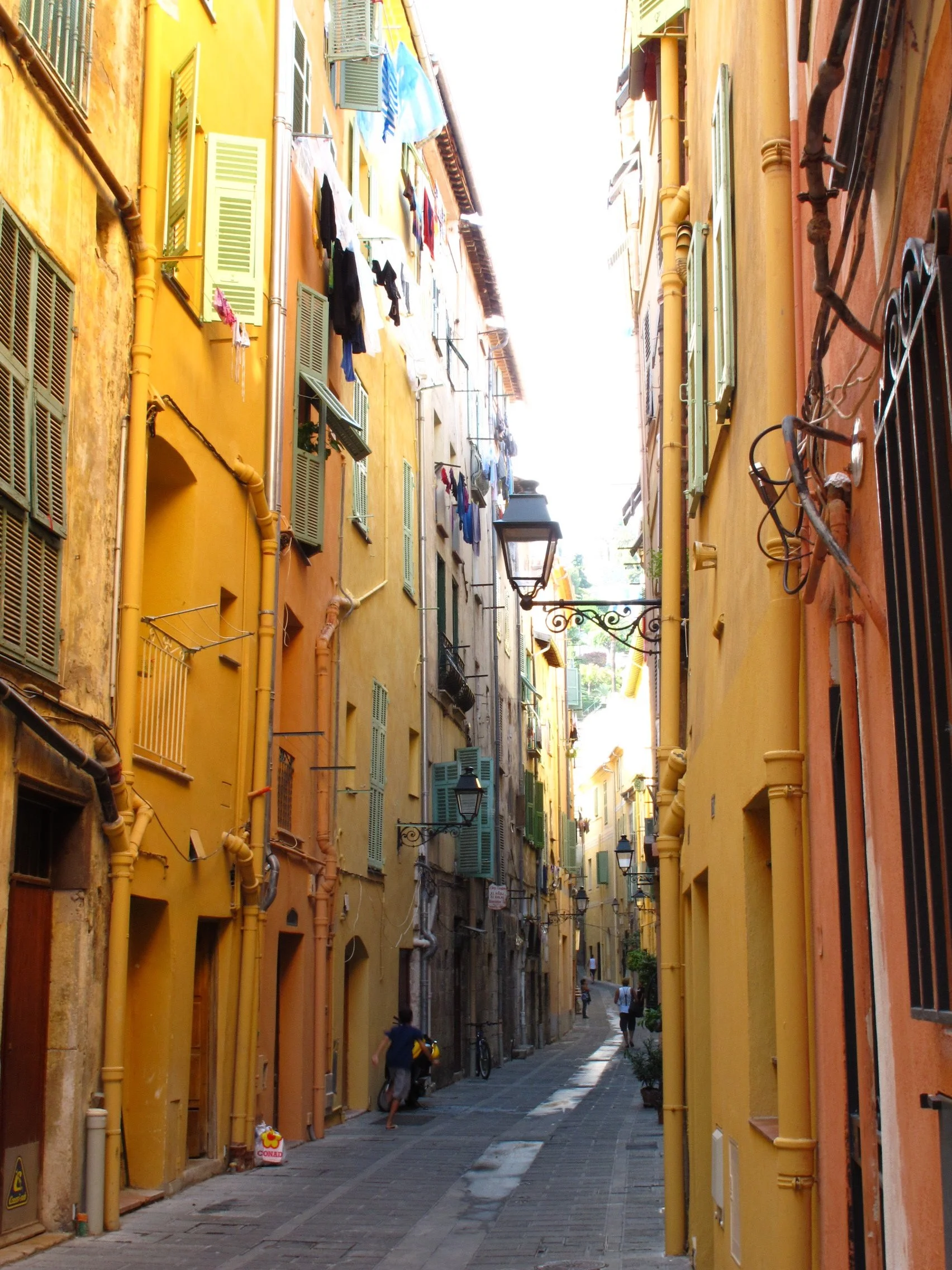 A narrow alleyway with colorful buildings on either side, featuring green shutters, laundry hanging, old street lamps, and a couple of people walking.