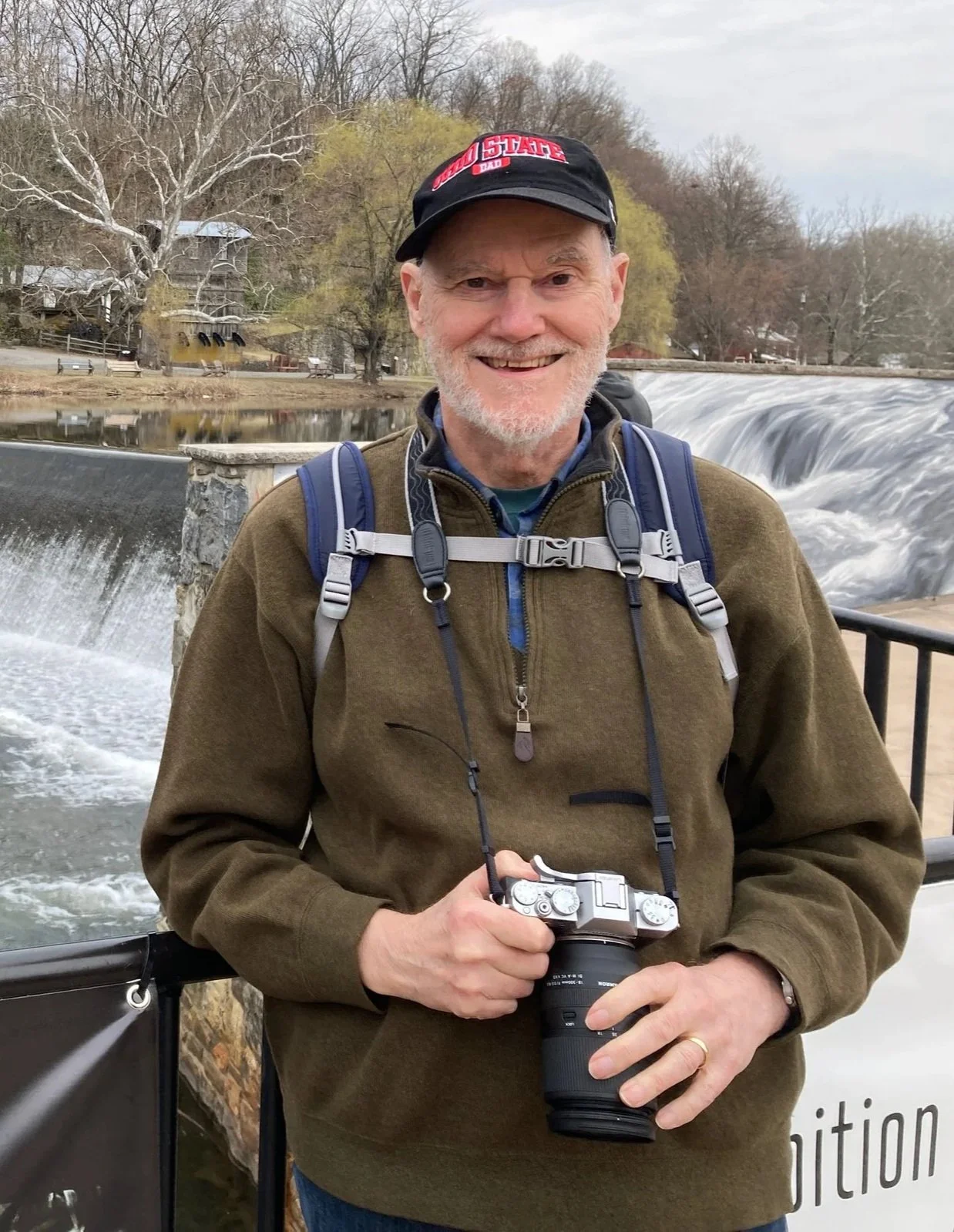 Smiling older man with a camera around his neck, standing outdoors near a waterfall, wearing a brown jacket, blue backpack, and a black cap with red text, with trees and a river in the background.