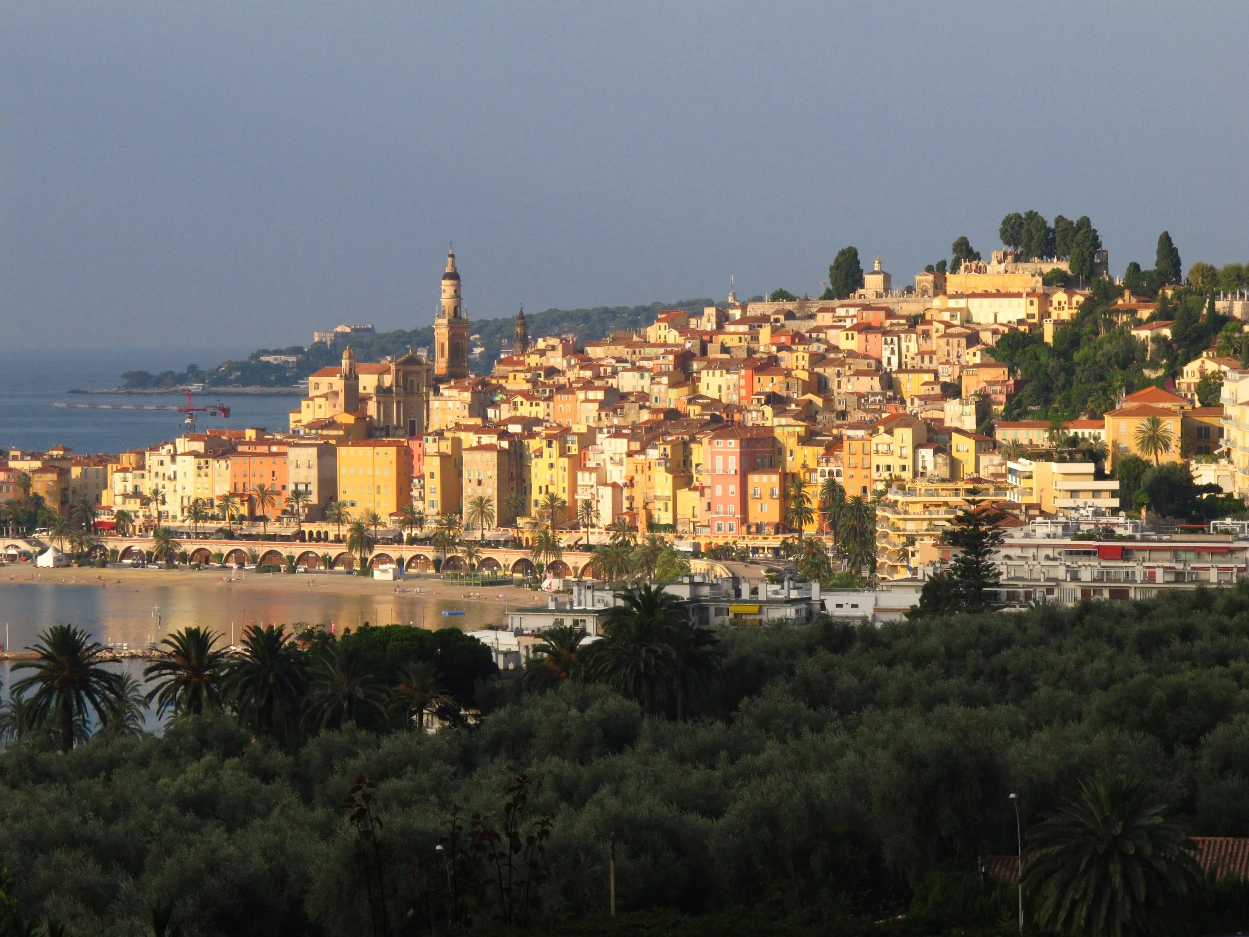 A coastal hillside town with colorful buildings, two prominent church towers, and a castle or fort at the top, overlooking the water and a sandy beach in the foreground.
