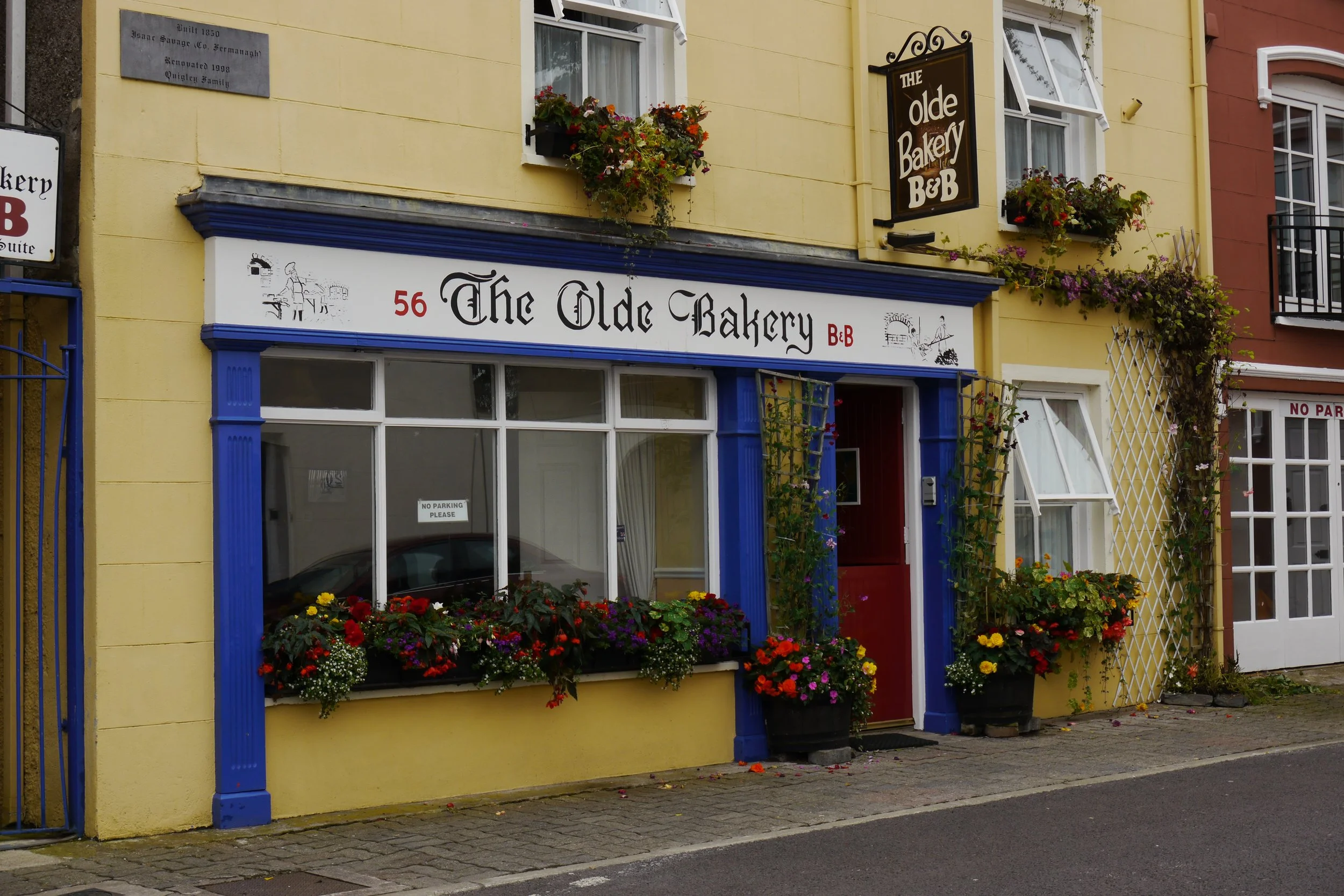 The yellow building houses a bakery called 'The Olde Bakery B&B' with a white sign accented in blue, and the number 56. There are colorful flowers in window boxes and planters along the sidewalk. A black sign on the building reads 'The Olde Bakery B&