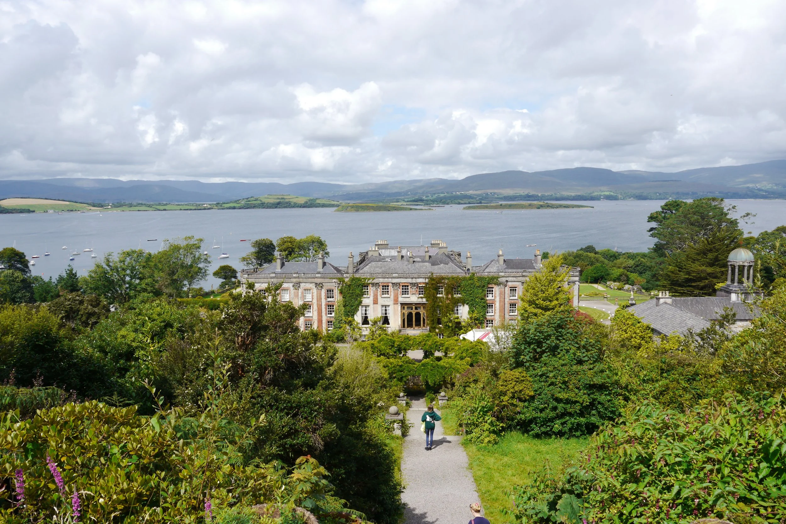 A scenic view of a large, historic building surrounded by lush green trees and gardens, with a lake and mountain range in the background. Two people are walking on a gravel path leading towards the building, and a dog is visible at the bottom of the 