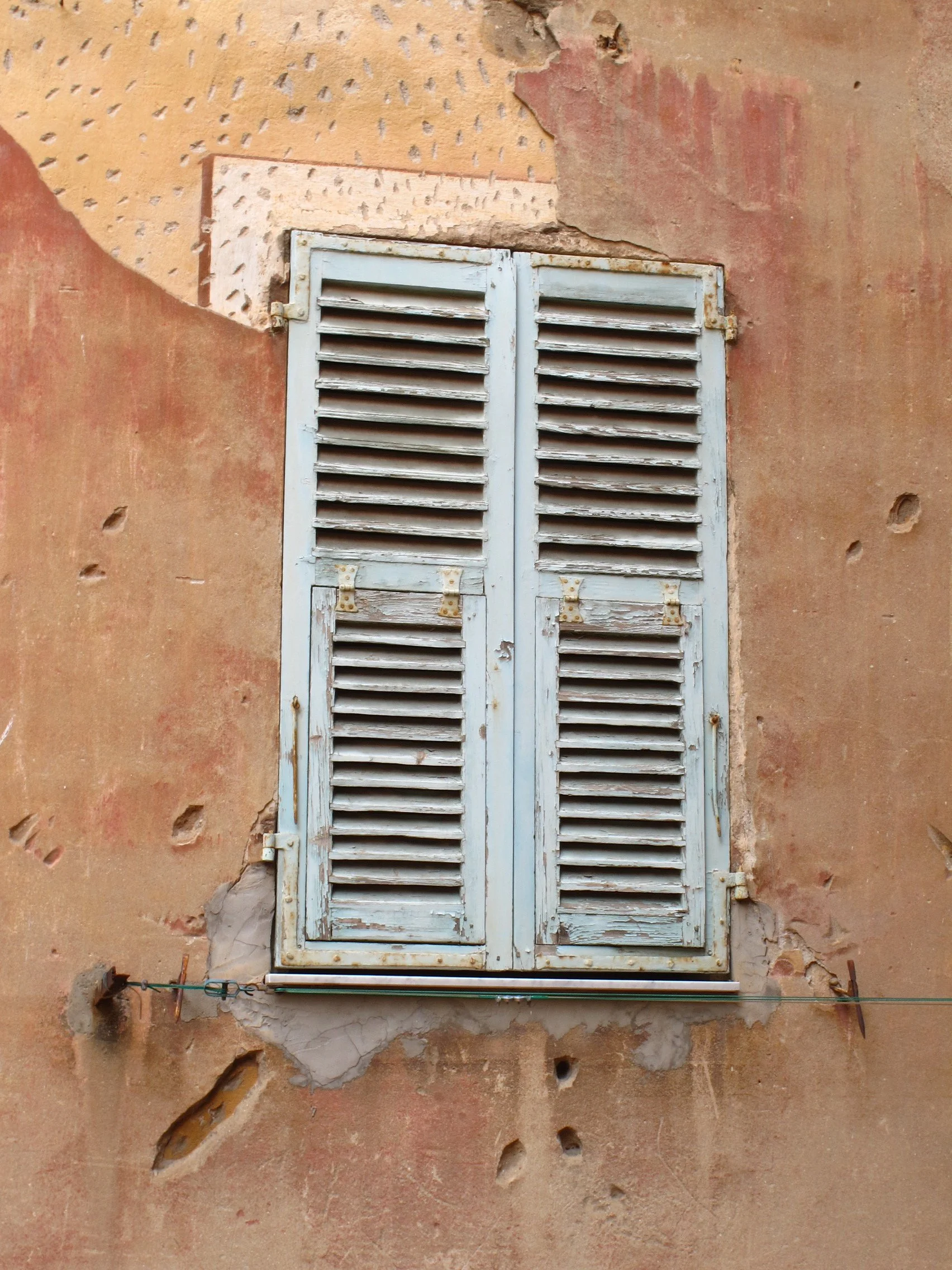 An old, weathered white window shutter attached to a deteriorating wall with peeling paint and holes.