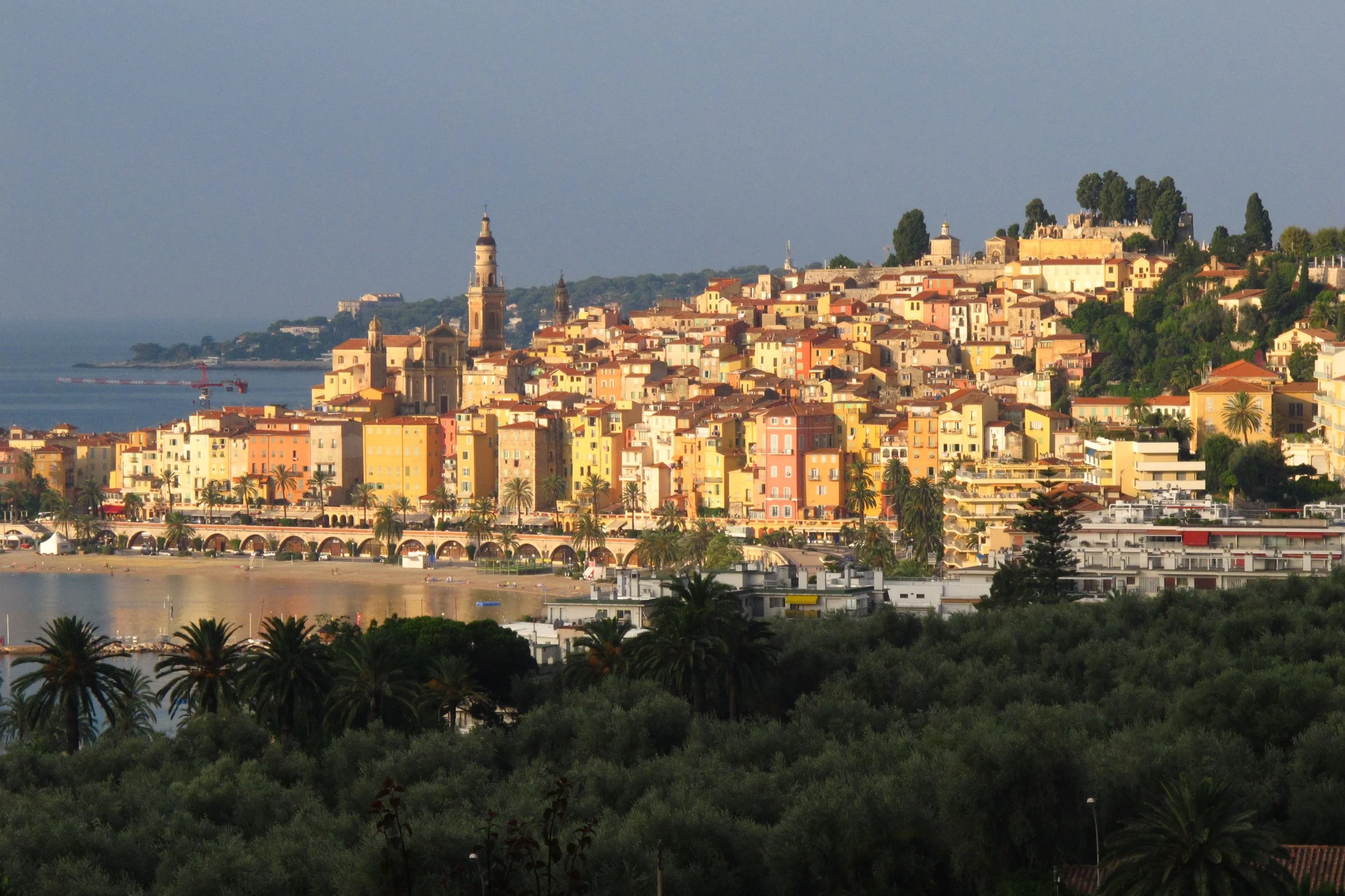 A coastal cityscape with colorful buildings on a hillside, a church with a tall bell tower, and a body of water in the foreground. Palms and lush greenery are also visible.