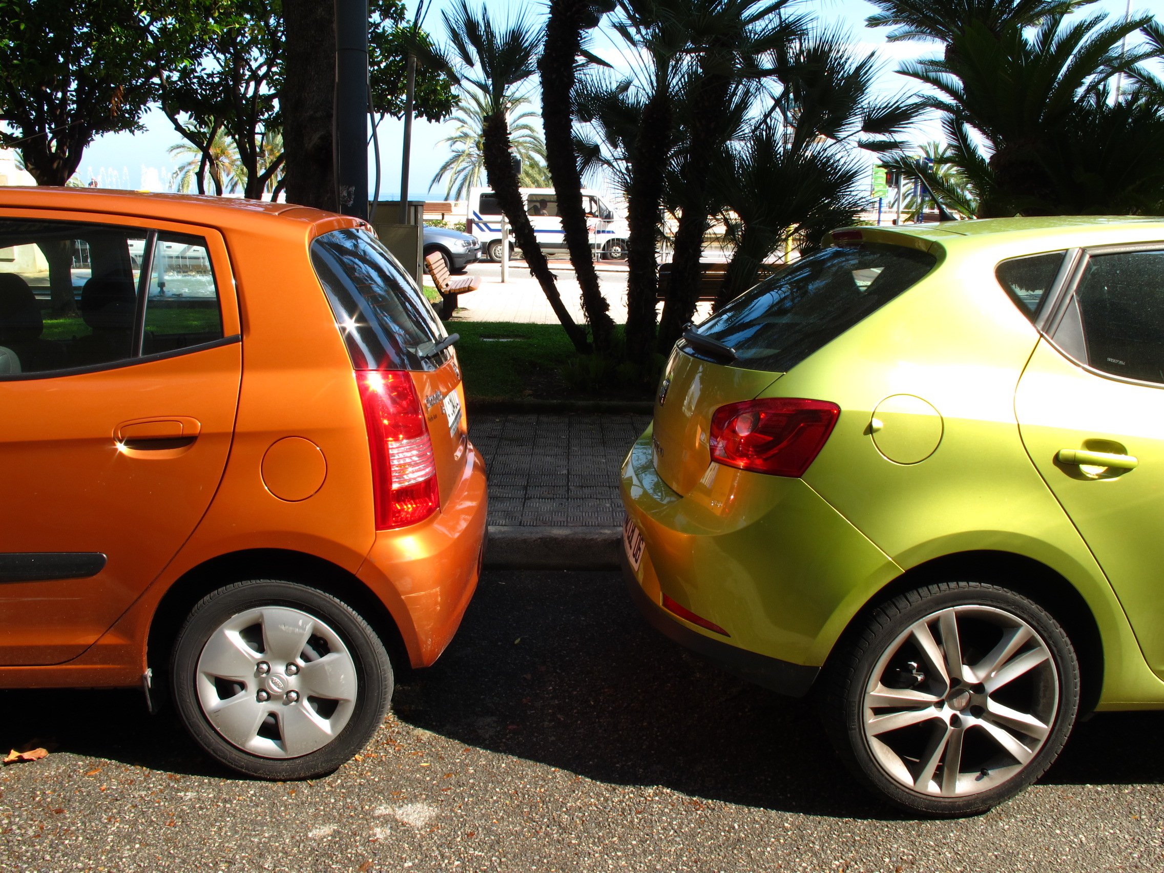Two parked cars, one orange and one yellow, on a gravel surface near palm trees and lush greenery with a view of the ocean in the background.