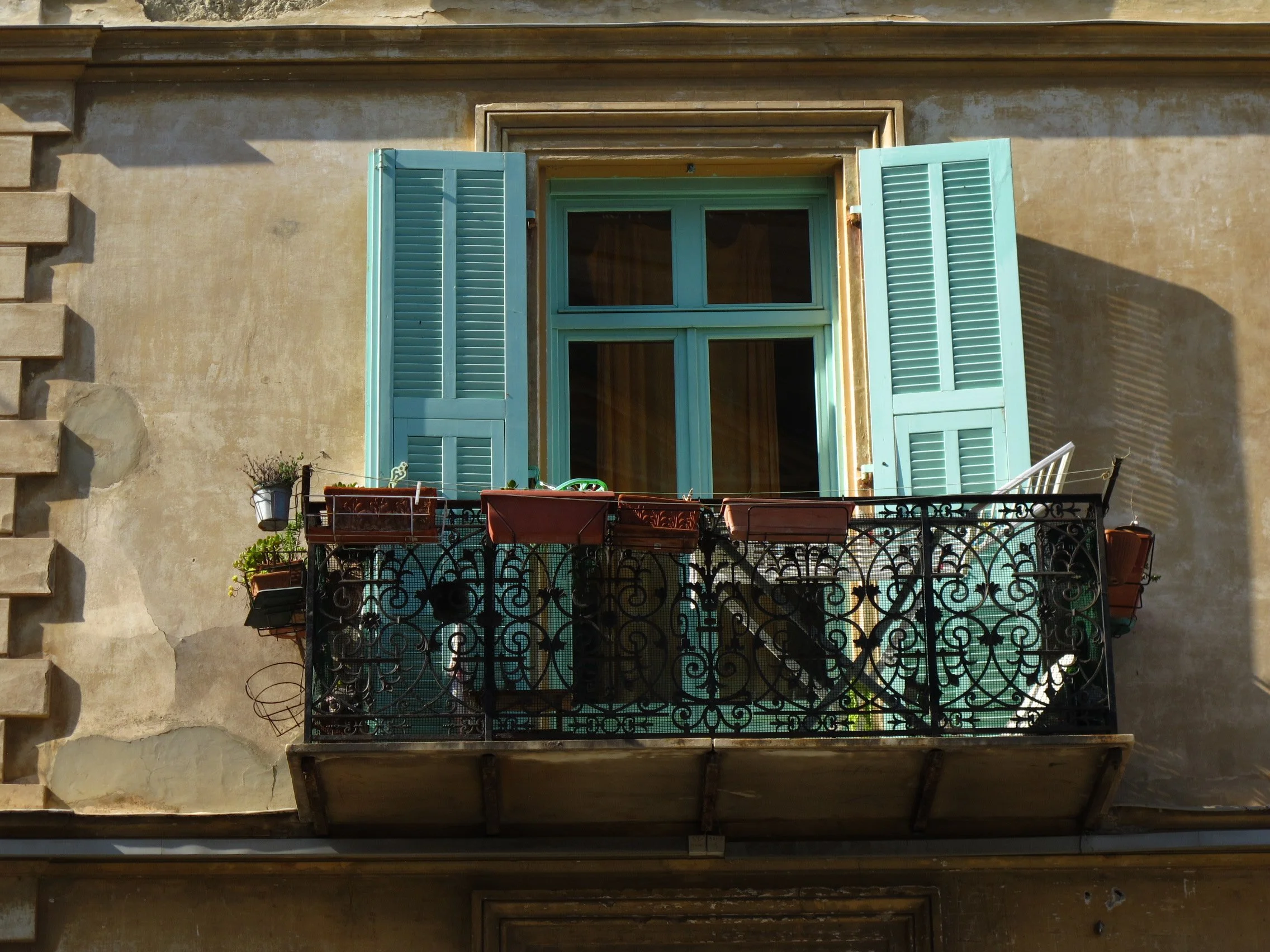 A balcony with potted plants and chairs, a window with teal shutters, and a wrought iron railing, viewed from above.