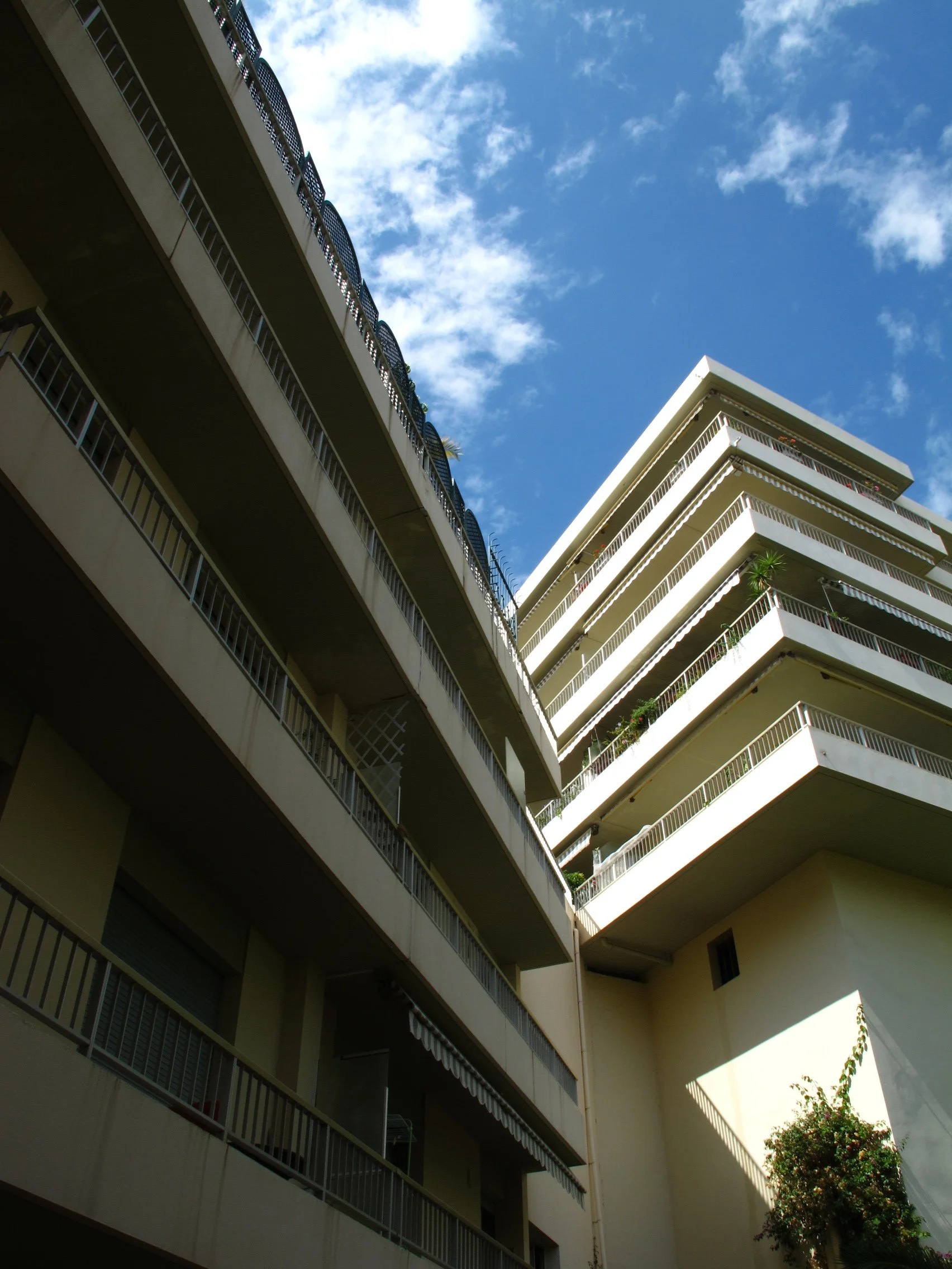 Multiple beige residential buildings with black railings and balconies under a partly cloudy blue sky, with trees and plants visible near the buildings.