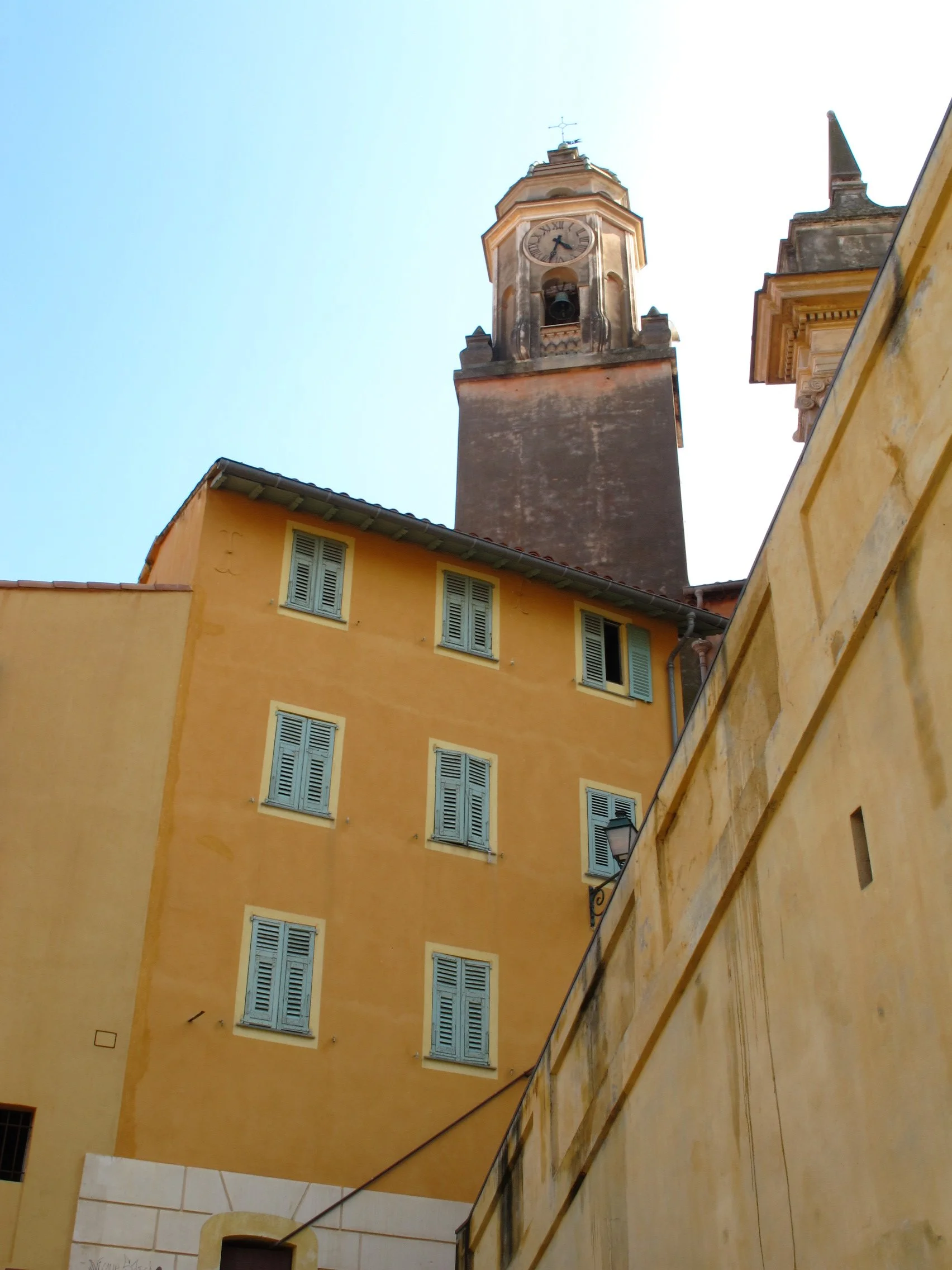 A tall clock tower with a clock face showing the time as approximately 4:41, located behind peach-colored buildings with light blue window shutters, set against a clear blue sky.