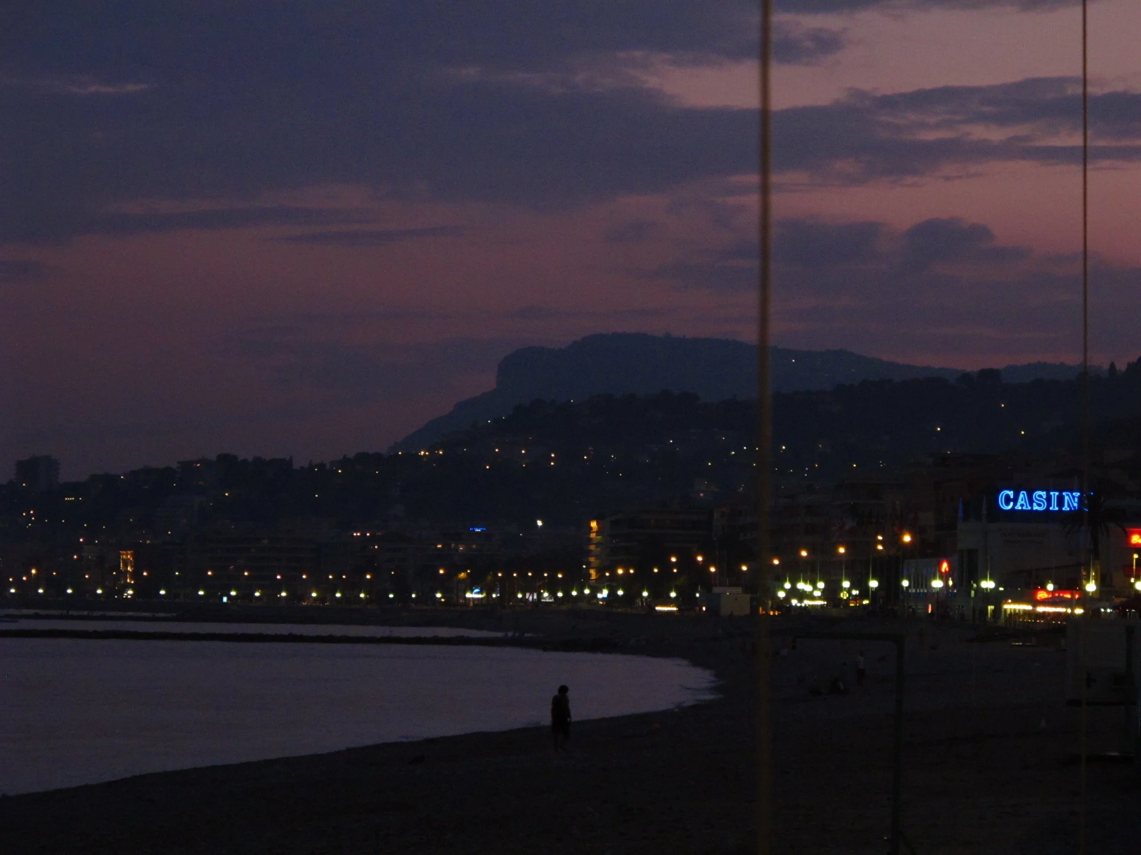 View of a cityscape at dusk with a mountain in the background and a large sign that reads 'CASIN' illuminated in blue, along with scattered lights and a silhouette of a person standing near the water.