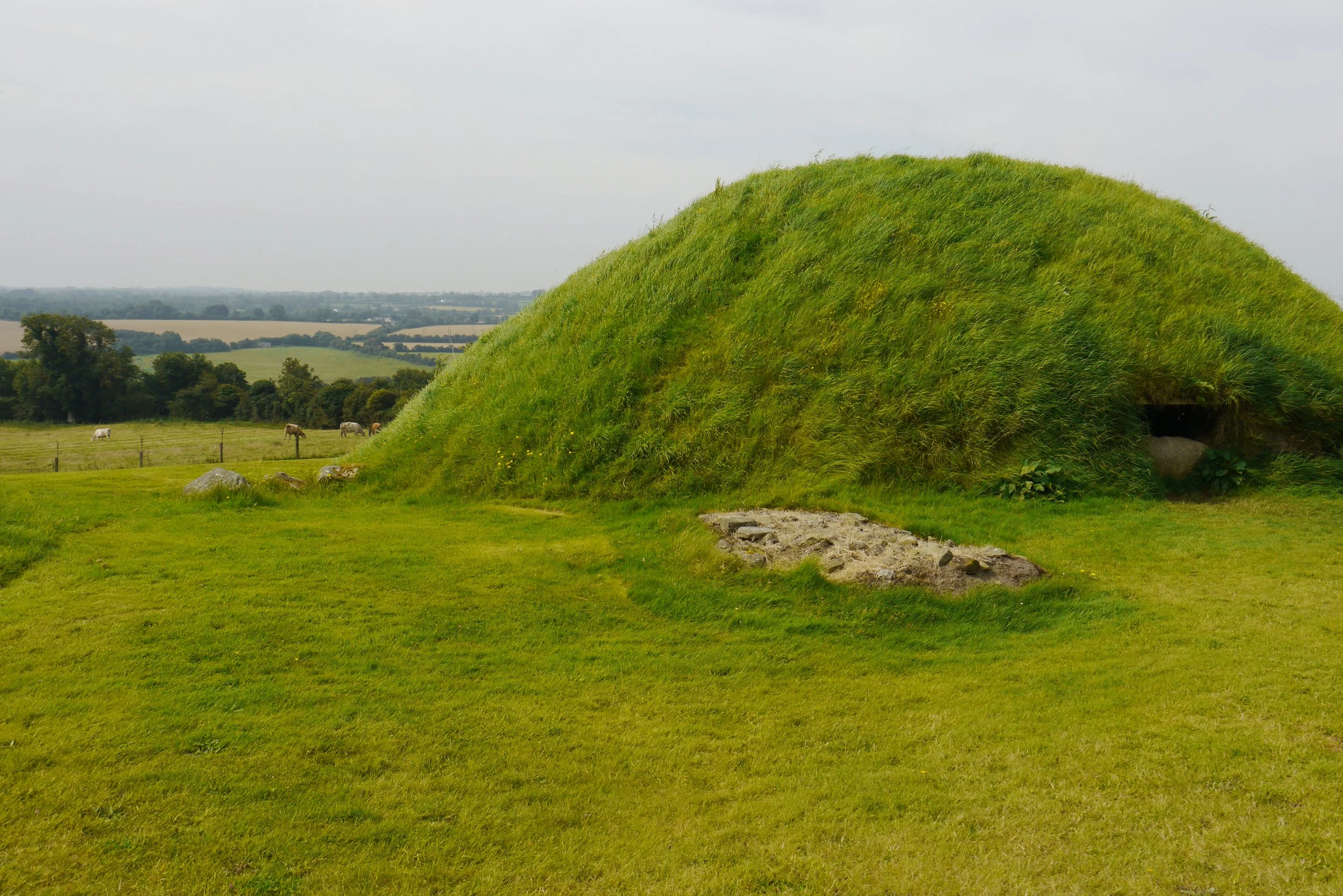 A large grassy mound with a small opening at its base, set in a rural landscape with fields, trees, and a cloudy sky in the background.