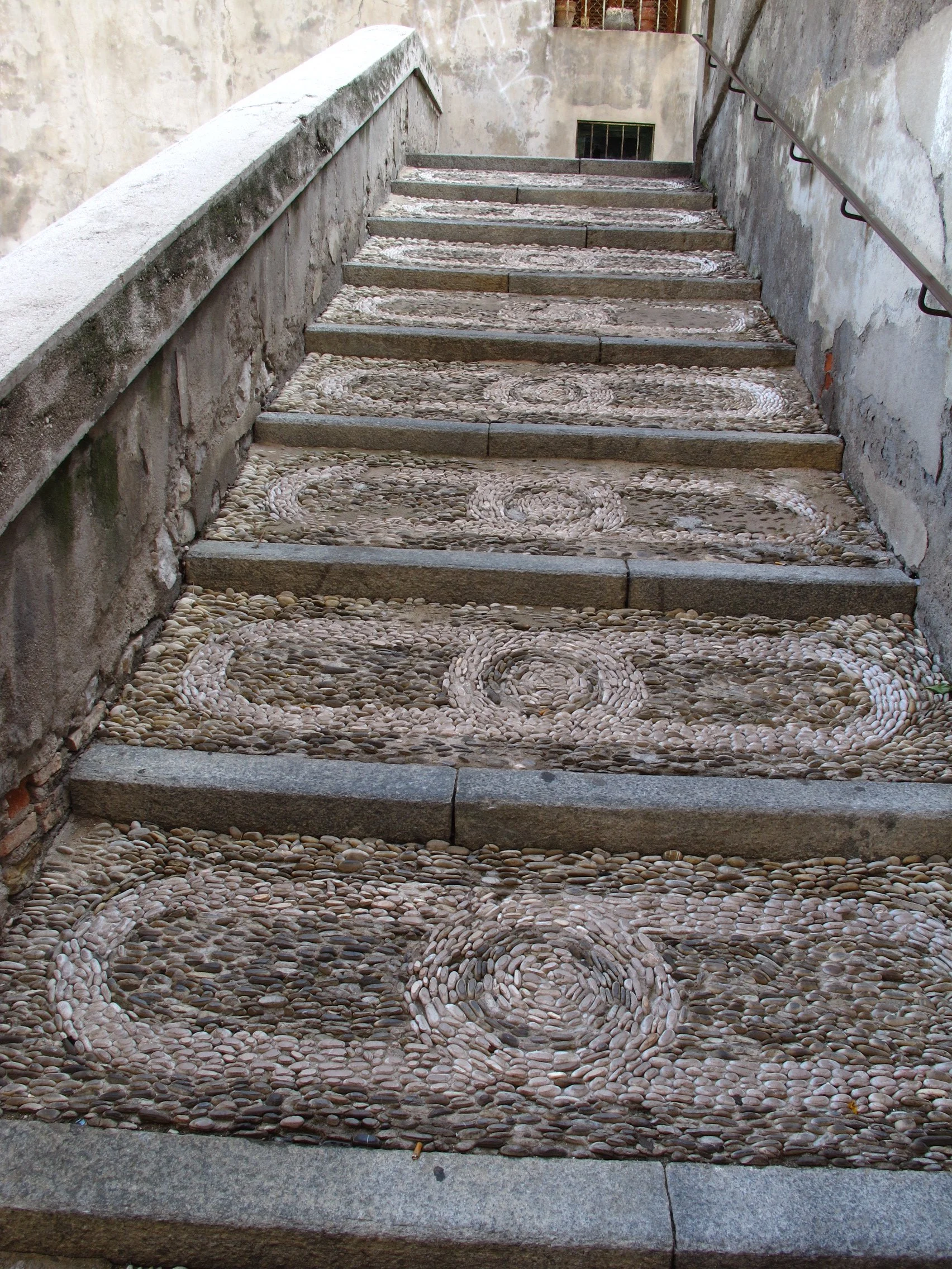 Stone stairs with decorative pebble patterns on each step, enclosed by rough plastered walls and a metal handrail.