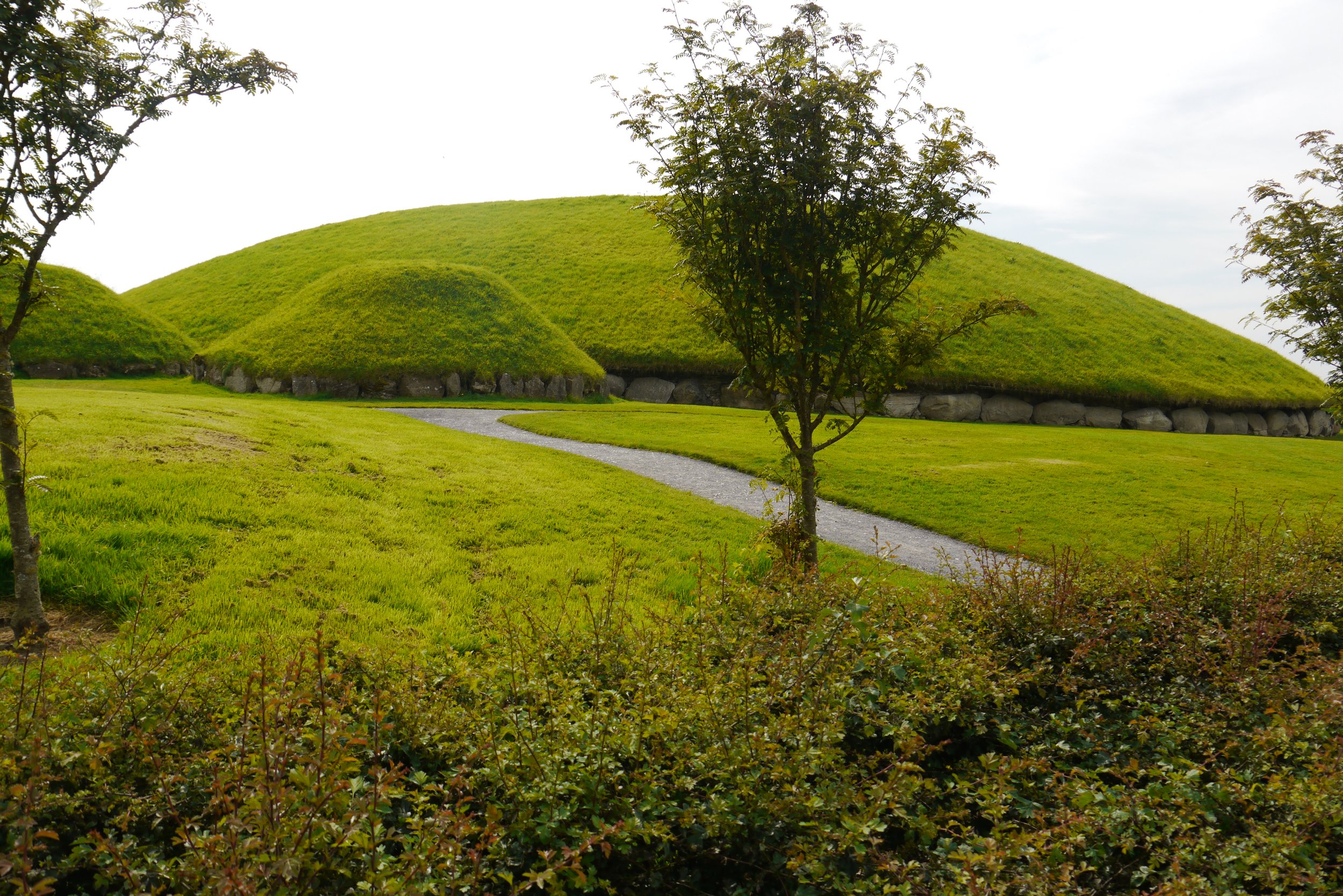 Green grassy landscape with small hills, a winding gravel path, and trees in the foreground, under a cloudy sky.