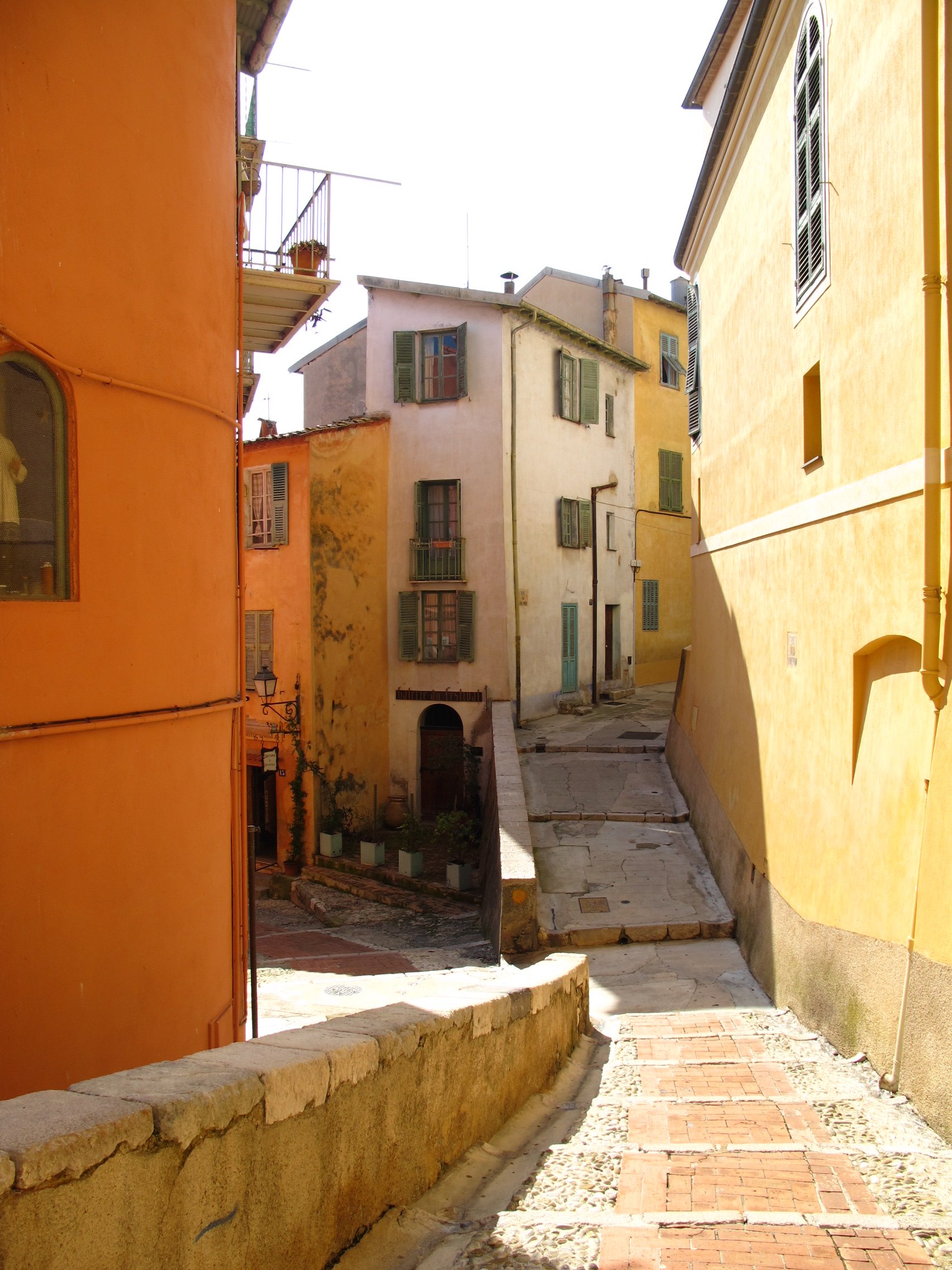 Narrow alleyway with colorful old buildings, yellow and pink, with green shutters, and stone pavement under bright sunlight.