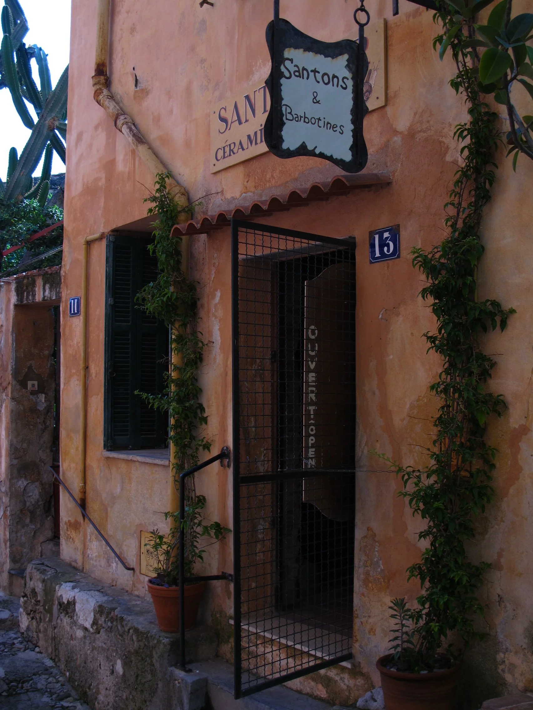 An exterior view of a building with a peach-colored stucco wall, green plants in pots, and an outdoor black metal cage. There are signs that read 'Santons & Barbottines' and 'SANTON CERAMIC.' The building has the numbers 11 and 15 on signs.