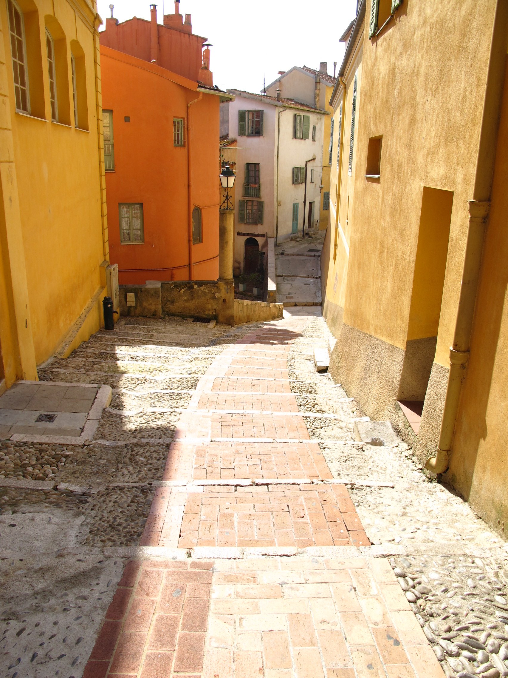 A narrow stone and brick staircase descending between colorful yellow and orange buildings on a sunny day in a European town.