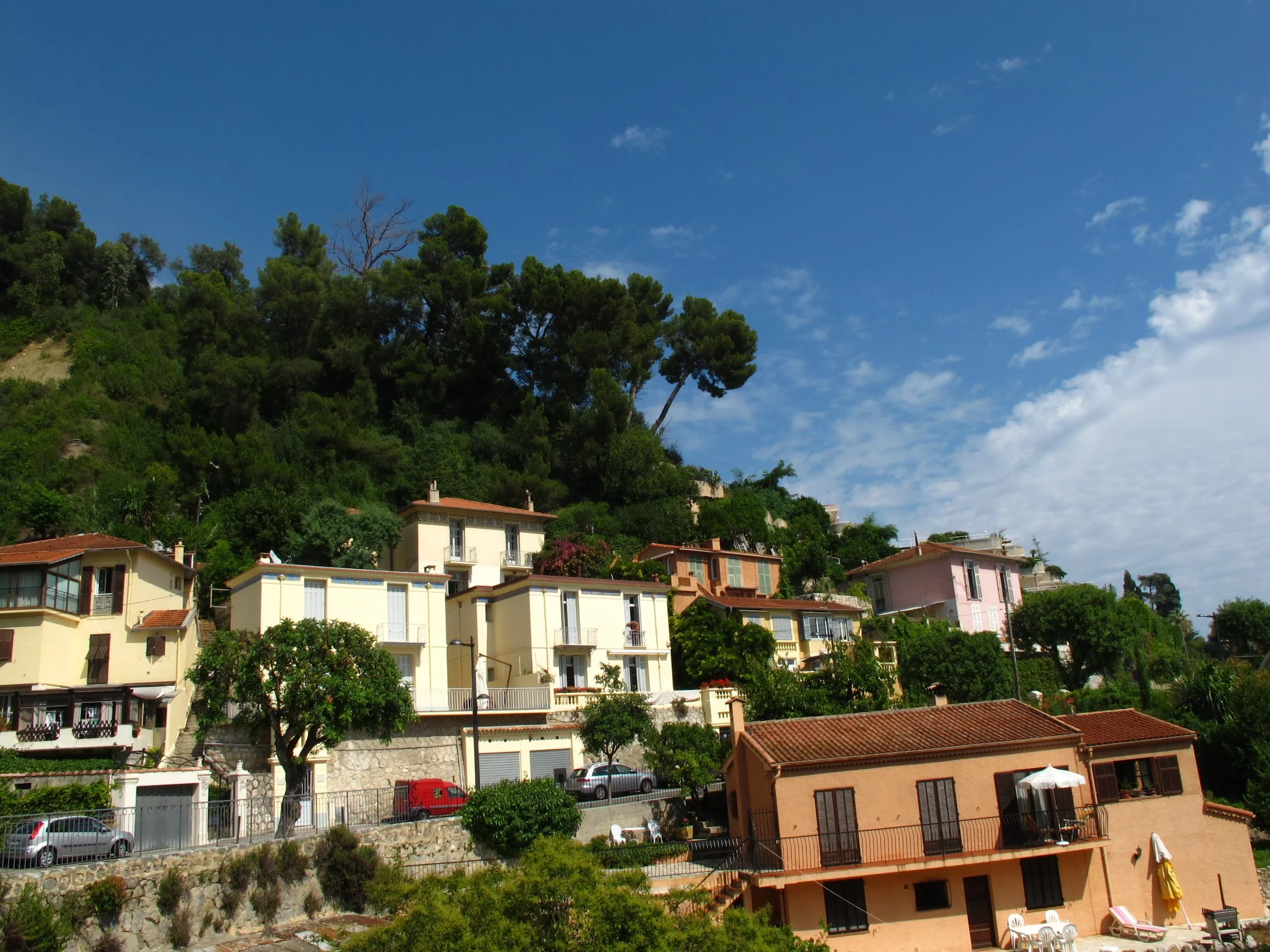 Houses on a hillside under a blue sky with scattered clouds, surrounded by trees.