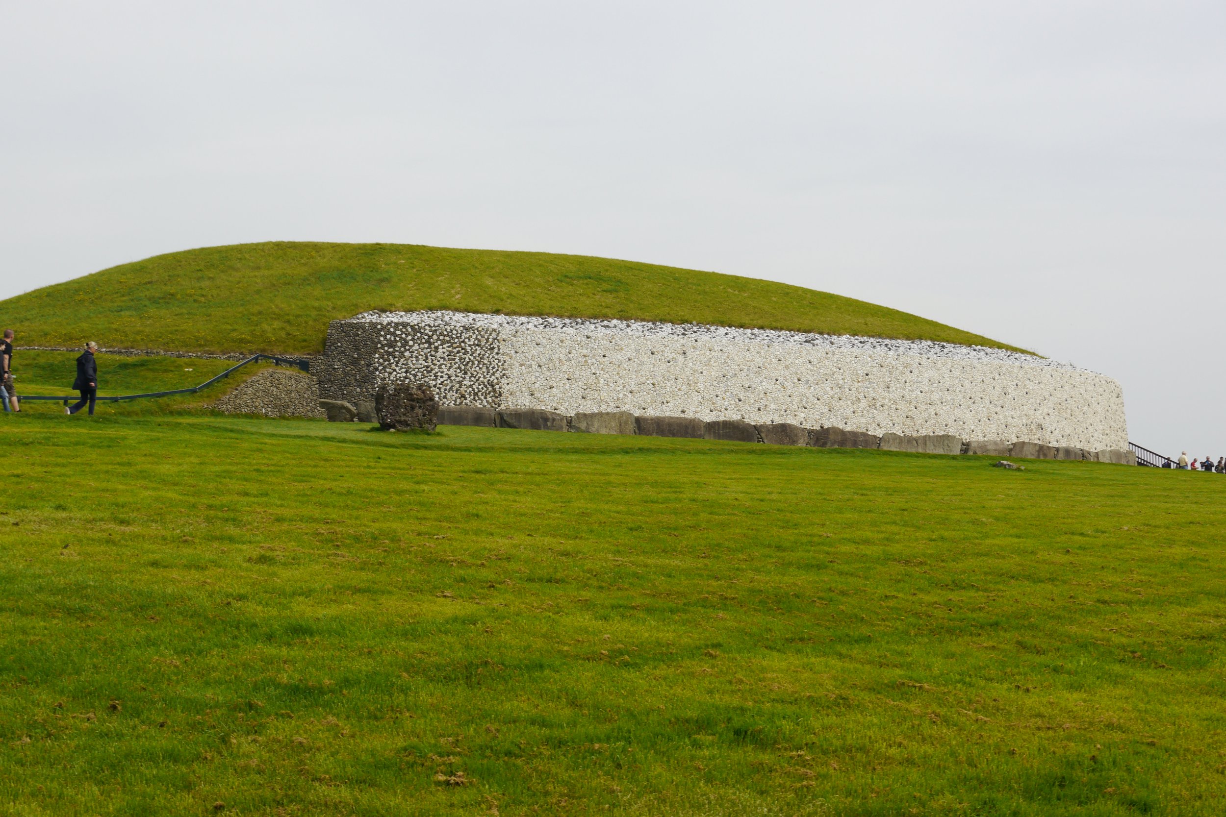 A grassy hill with a stone-and-cement foundation at the base, and people walking along a path nearby, under a cloudy sky.