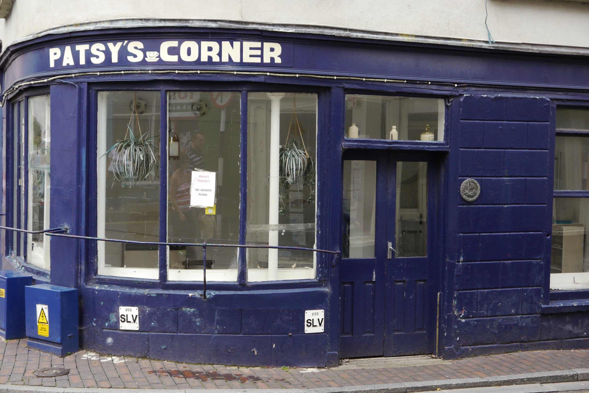 A blue-painted storefront named Patsy's Corner with large glass windows, a sign with white letters, and hanging plants inside. The store has a postal box and warning signs on the exterior.