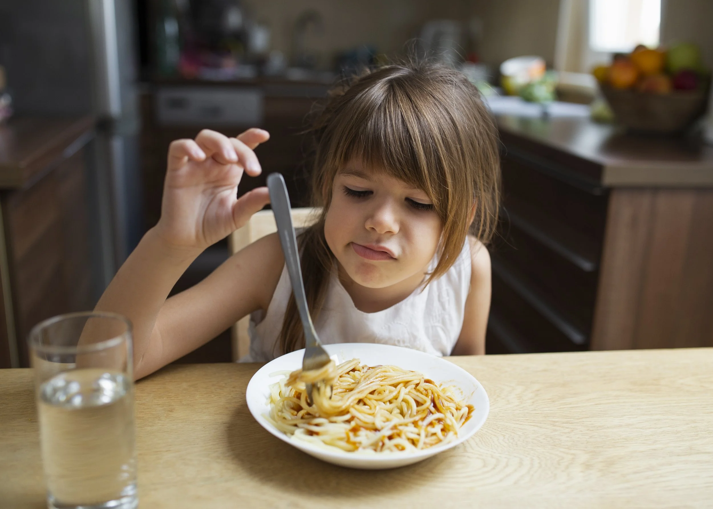 A young girl with brown hair and a white sleeveless top sitting at a wooden dining table, holding a fork with spaghetti, with a bowl of spaghetti in front of her. There are two glasses of water on the table and a blurred background of a kitchen with fruit on the counter.