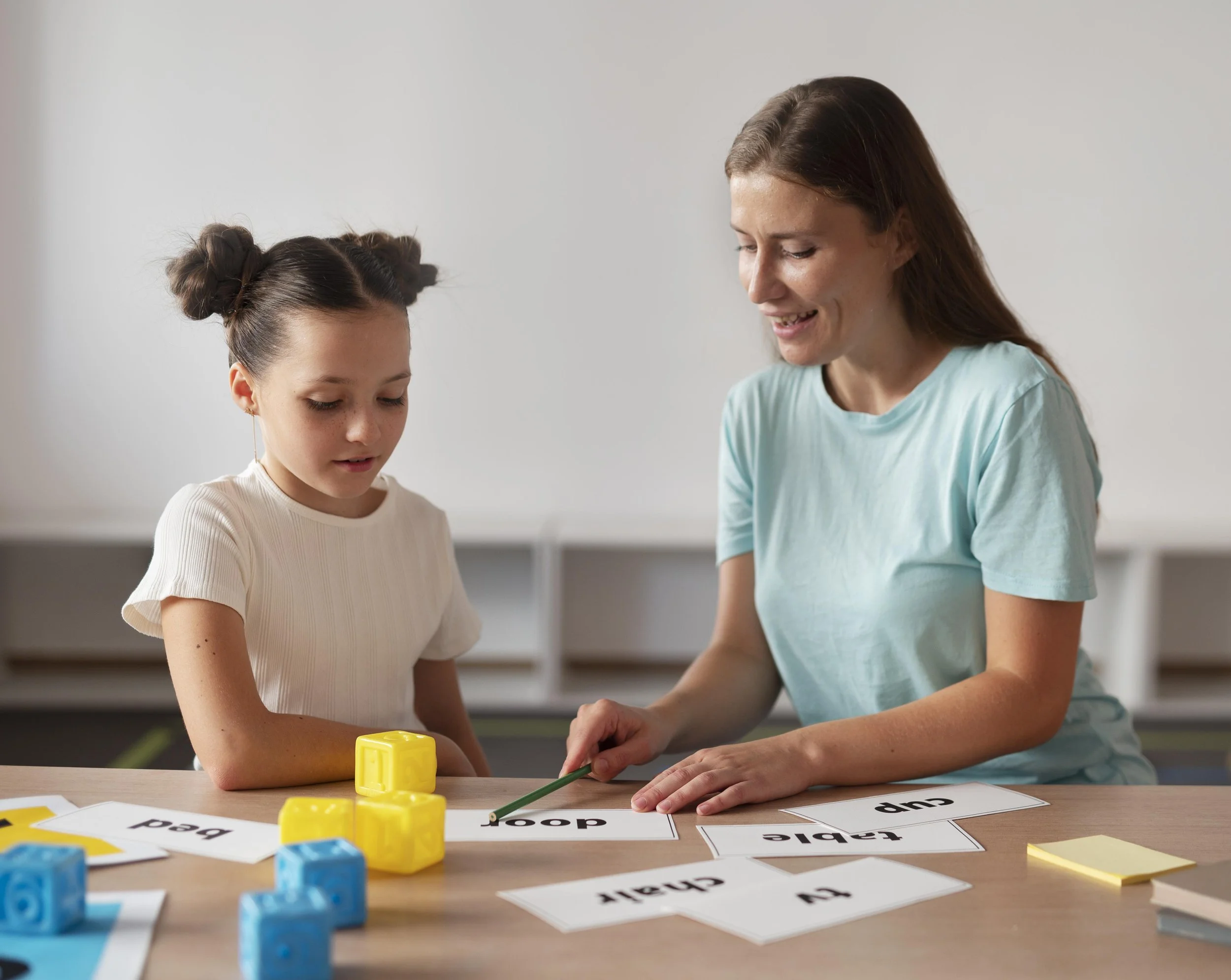 A young girl and an adult woman working together on an educational activity with word and picture cards on a wooden table. The girl has dark hair styled in two buns and is focused on the task. The woman has long brown hair and is smiling as she assists. Colorful plastic cubes and some word cards are on the table.